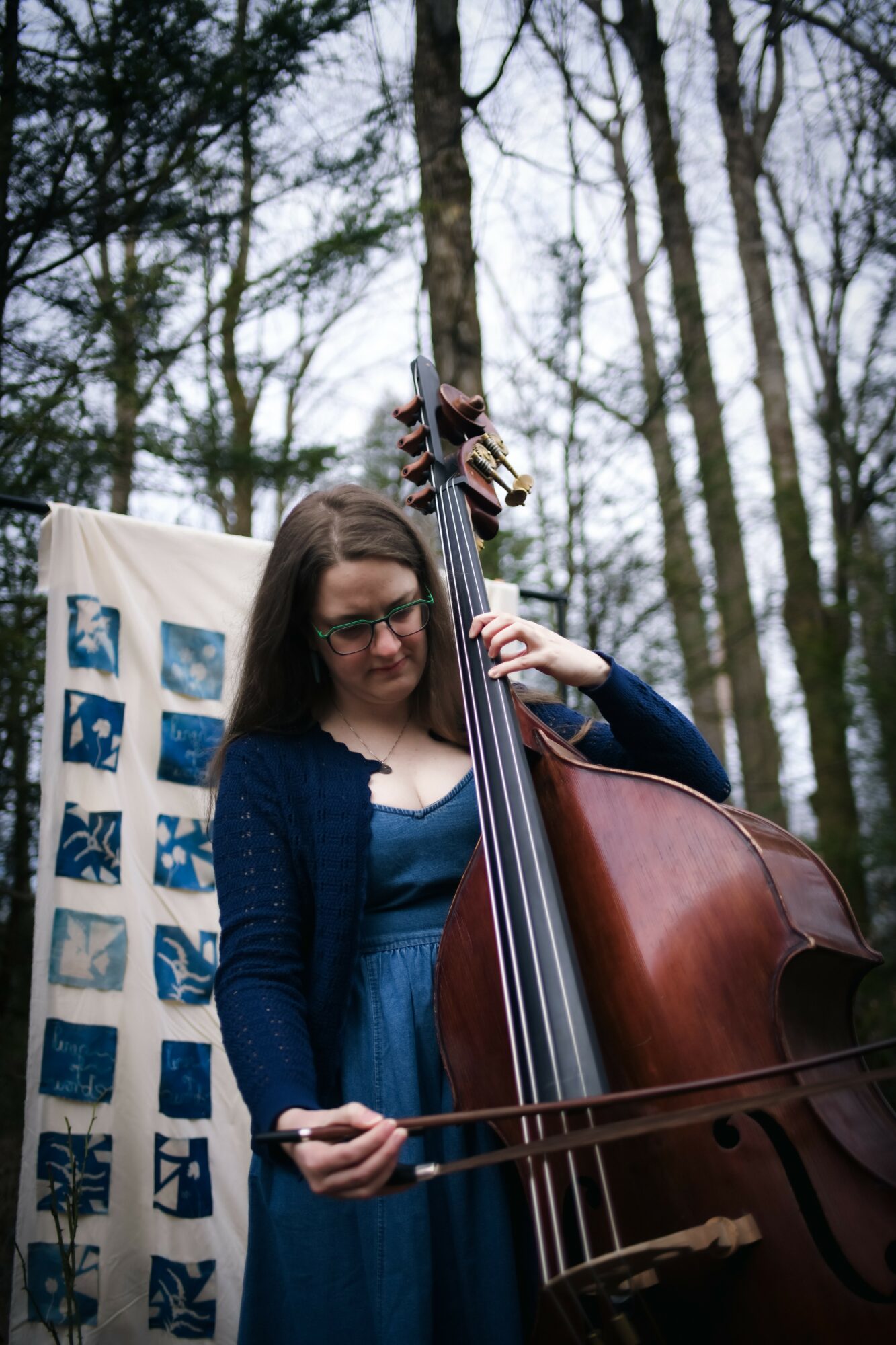 Woman playing double bass outdoors with trees in background and a patterned cloth nearby.