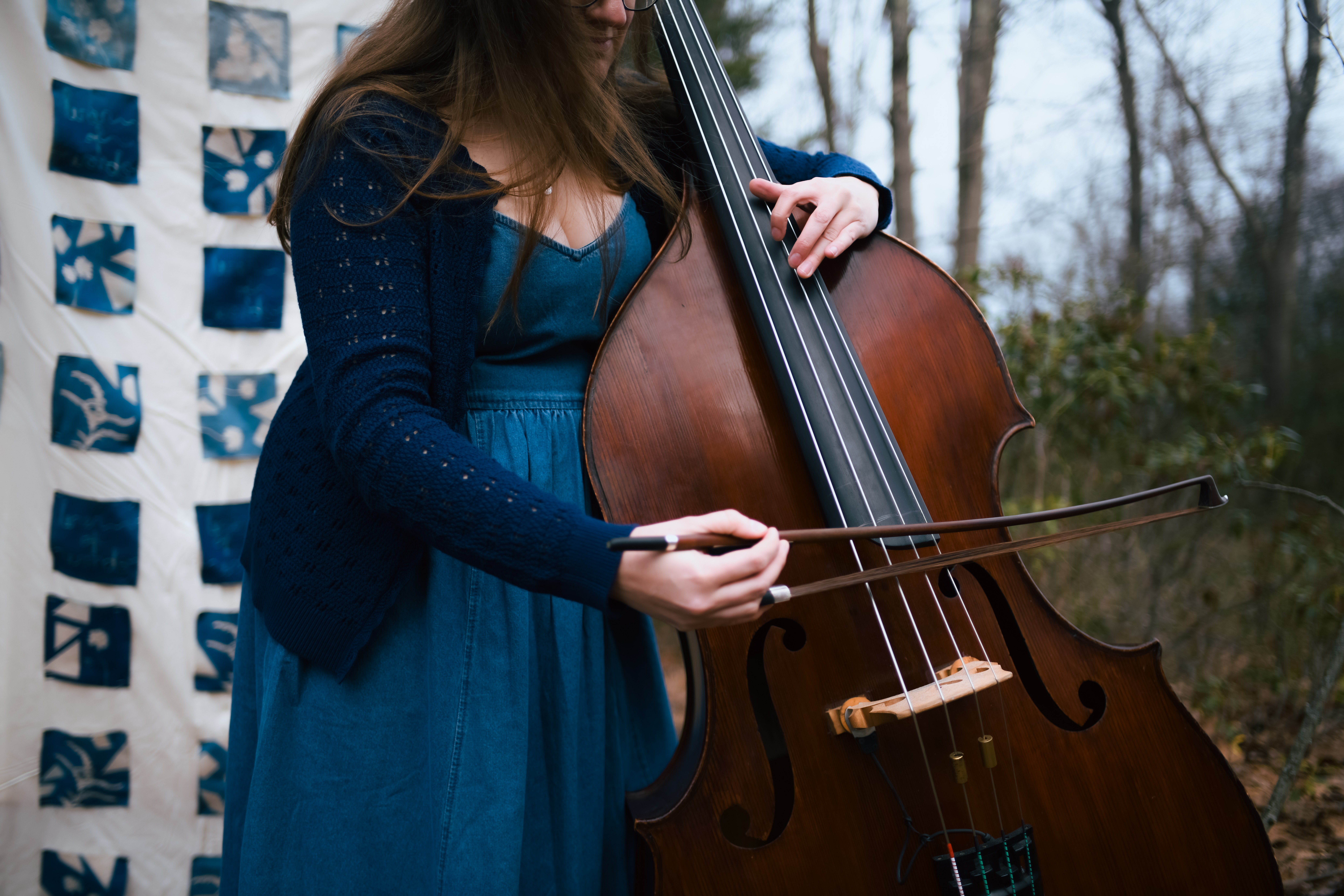 Person playing a double bass outdoors, wearing a blue dress and a dark cardigan, with trees in the background.