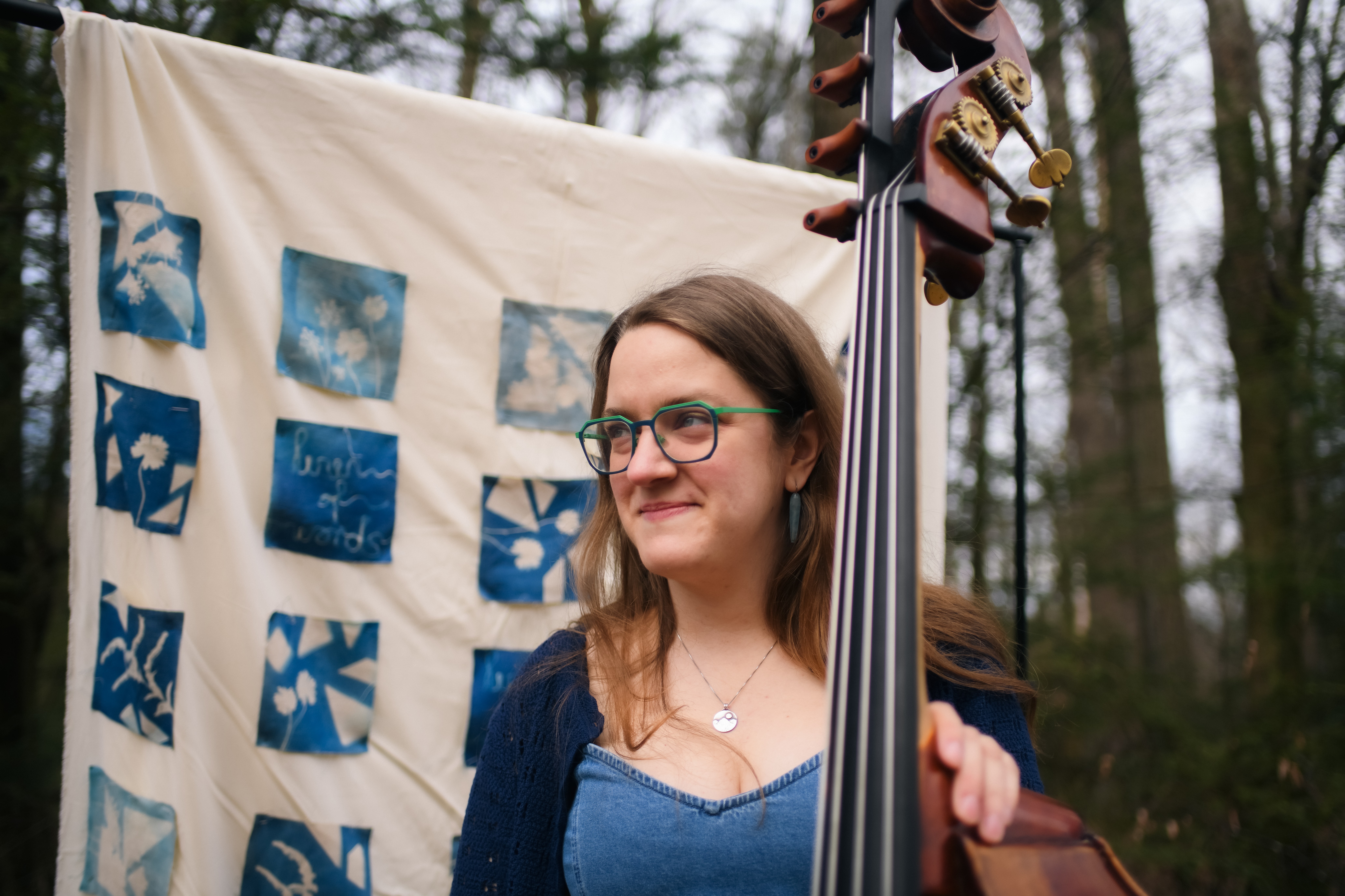 Young woman with glasses and long hair standing outdoors near a display of fabric squares, holding a musical instrument.