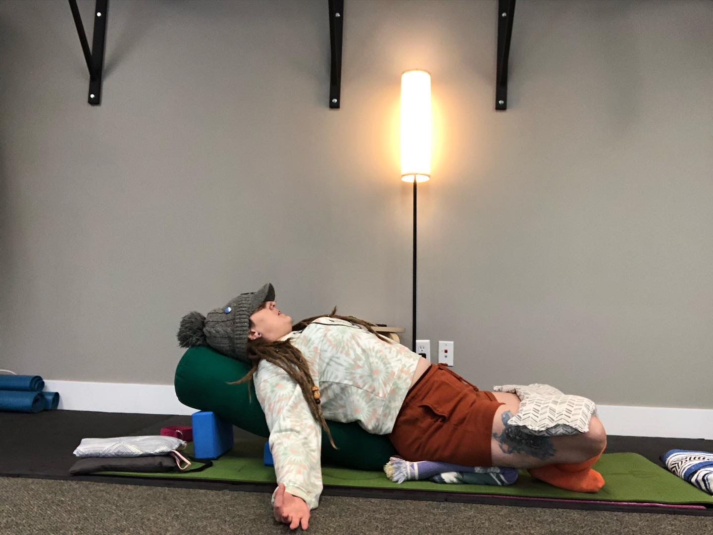 Person lying on side on yoga mat, head supported by cushion, in a room with wall-mounted shelves and a standing lamp.