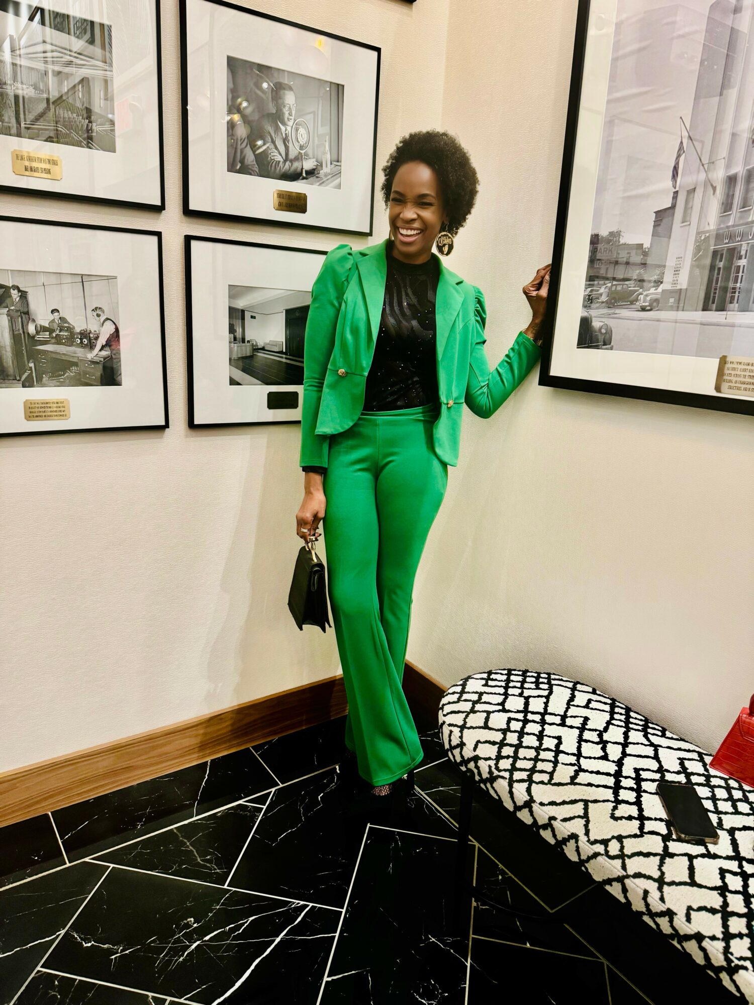 Woman in green pants and blazer standing in art gallery, smiling, holding a bag, next to framed pictures on wall.