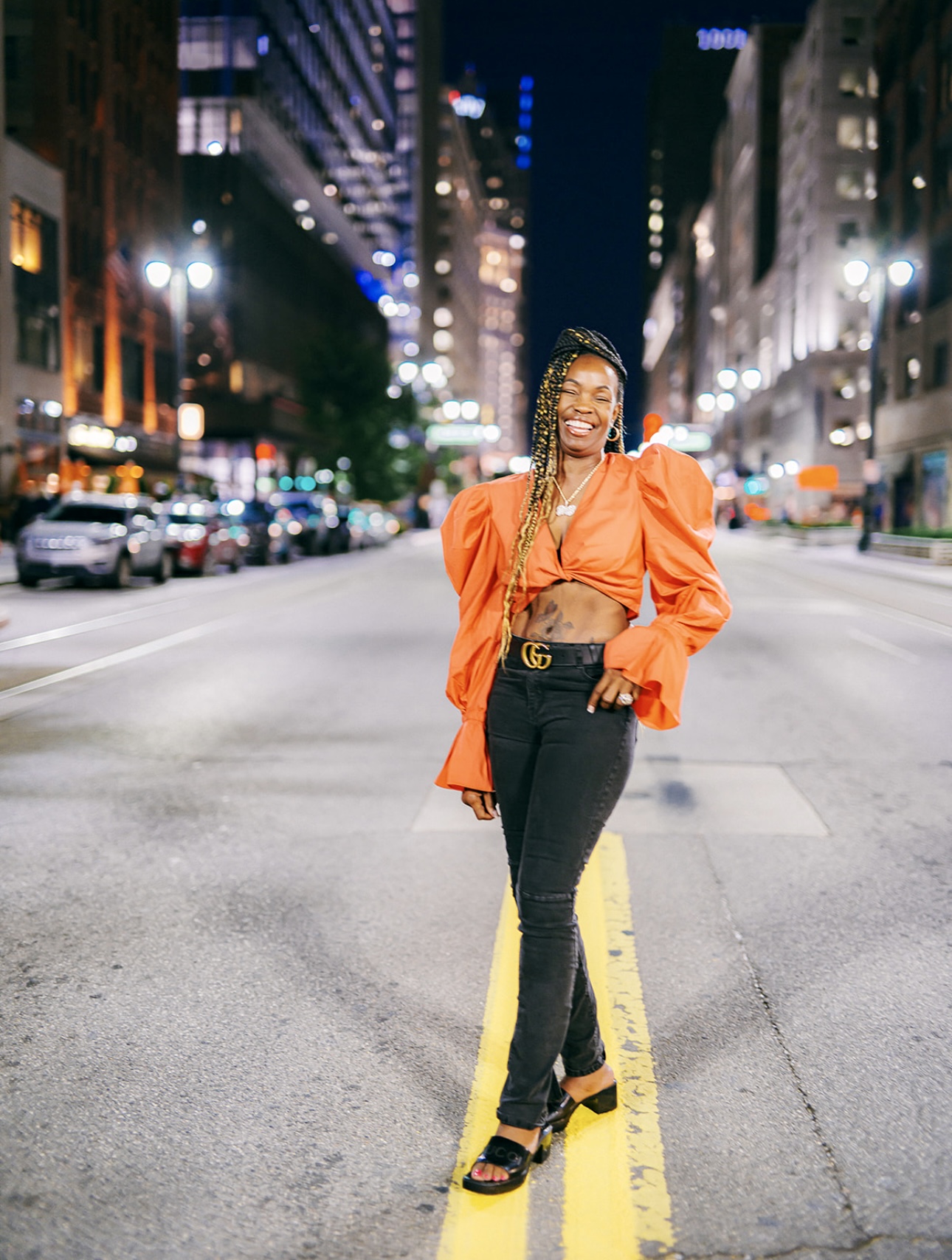 Woman standing in the middle of a city street at night, smiling, wearing an orange jacket and black pants.