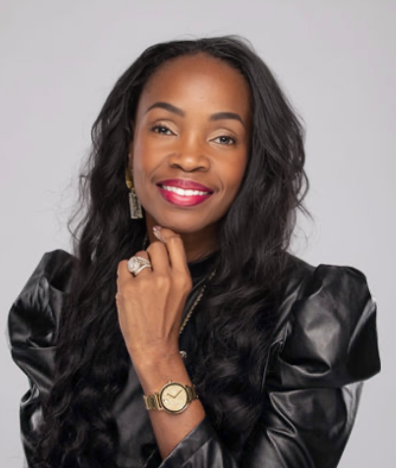 Woman with long wavy hair, smiling, wearing a black leather jacket, gold watch, earrings, and a ring, against a light background.