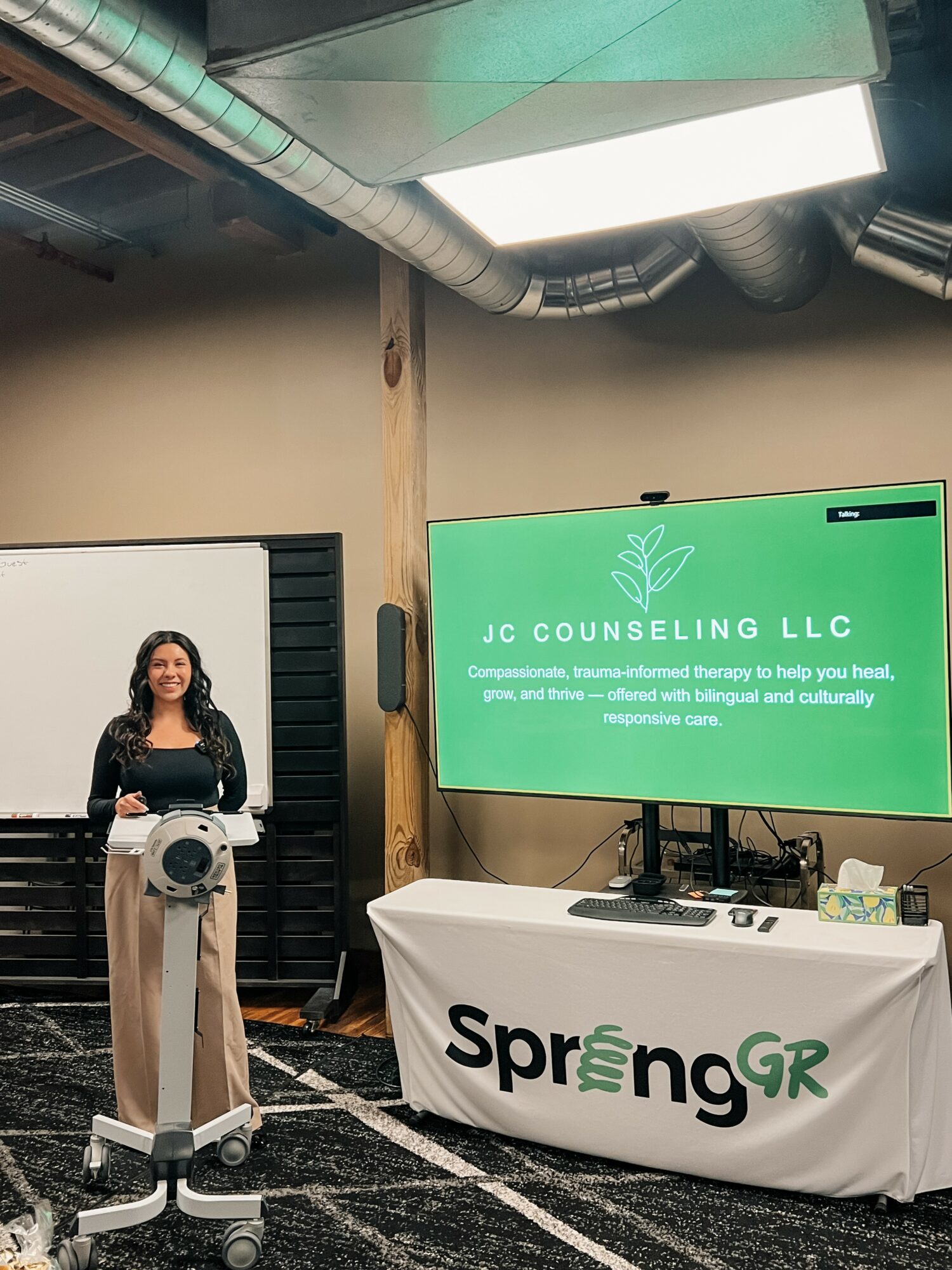 Woman standing next to a robot in a room with a large screen displaying 'JC Counseling LLC' and a table with a banner reading 'SpringGR'.