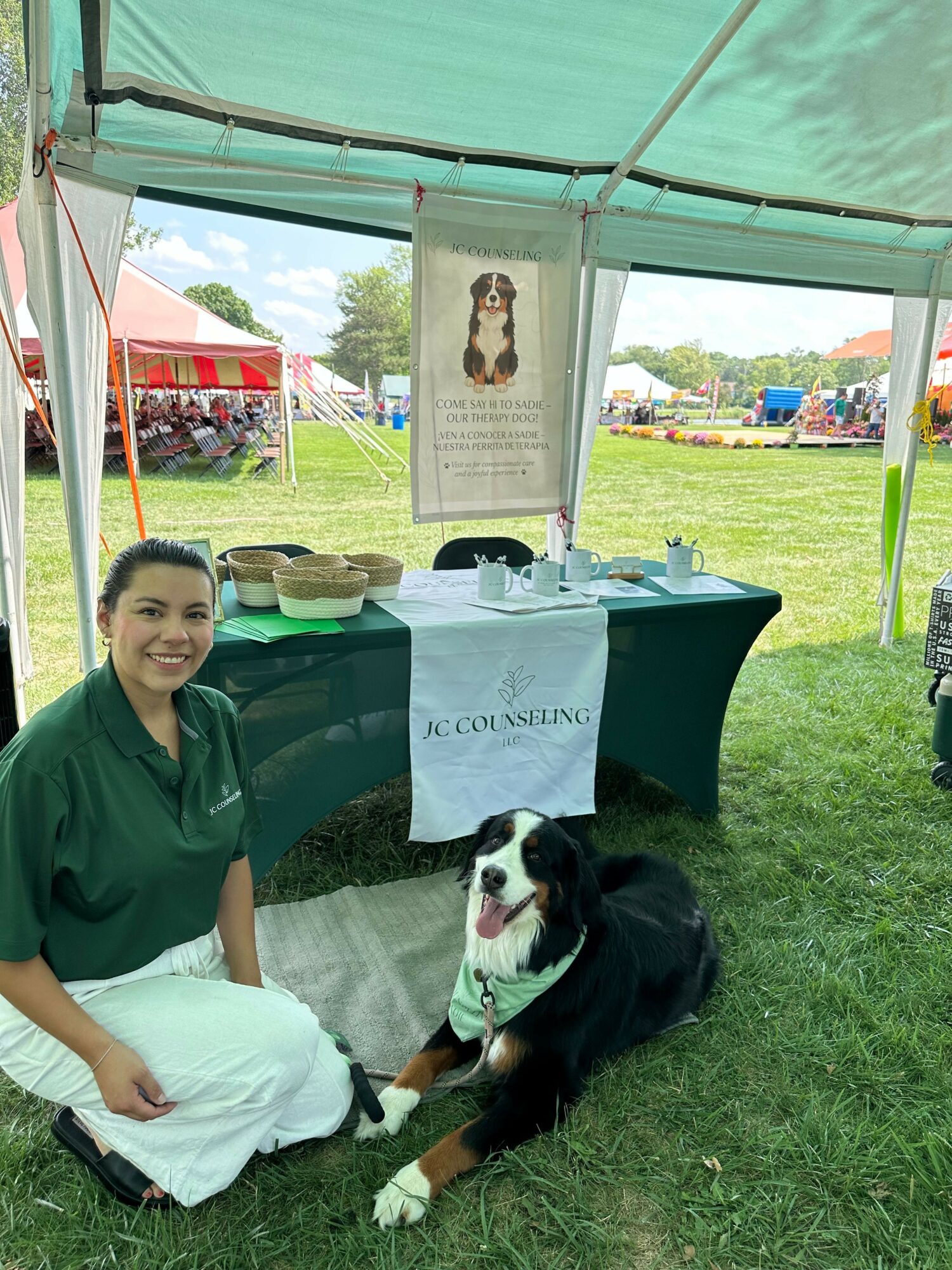 Woman sitting on grass with black and white dog under a tent at outdoor event, table with bowls and banner behind.