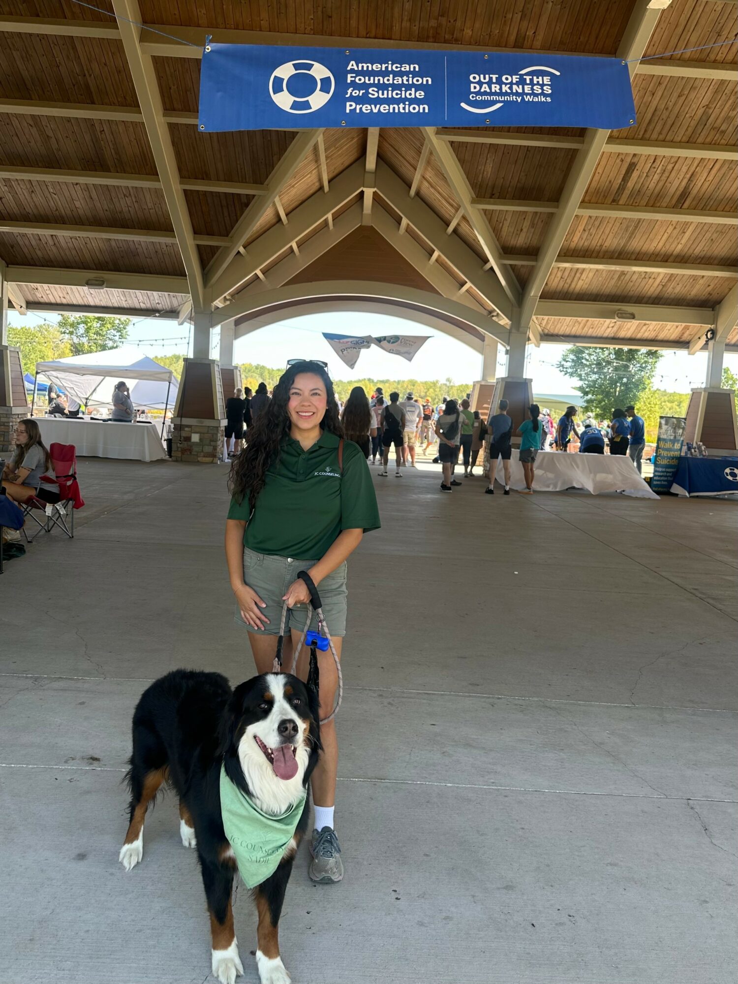 Woman standing with a black and white dog wearing a green bandana under a pavilion at an outdoor event.