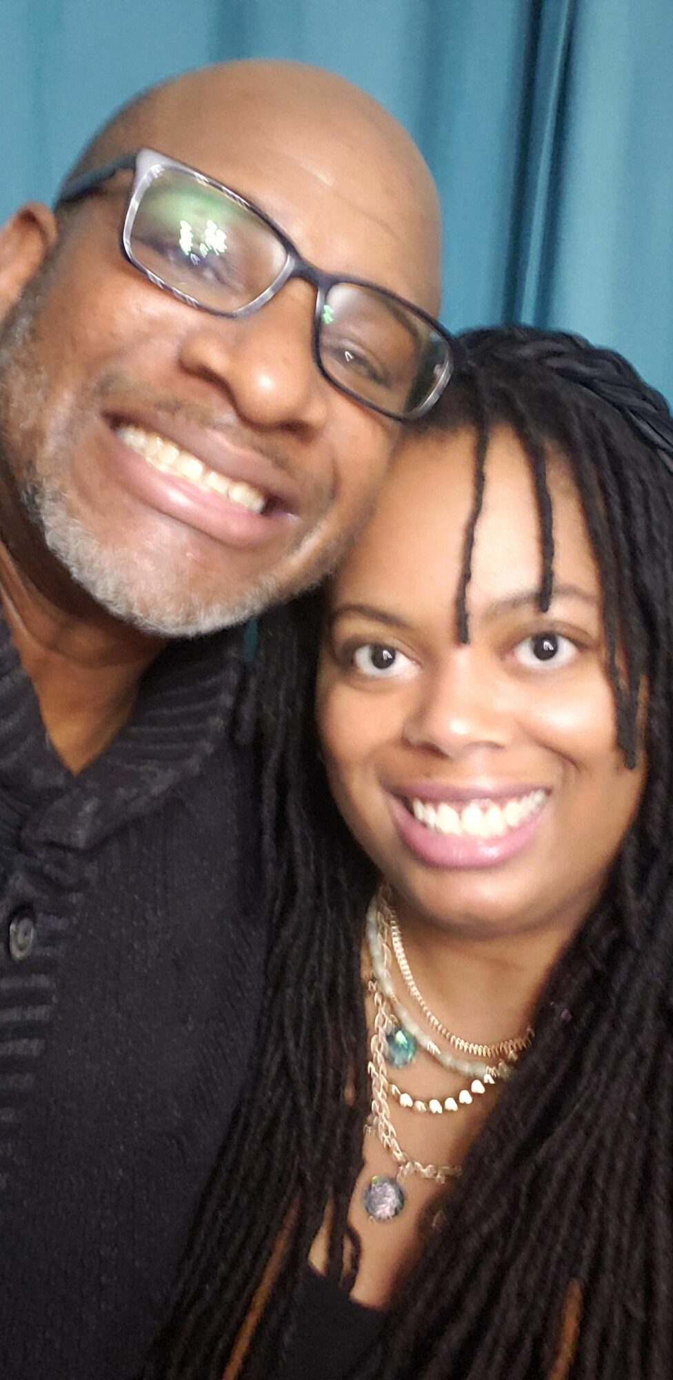Two smiling people close together, one wearing glasses and the other with long dreadlocks, in front of a blue curtain.