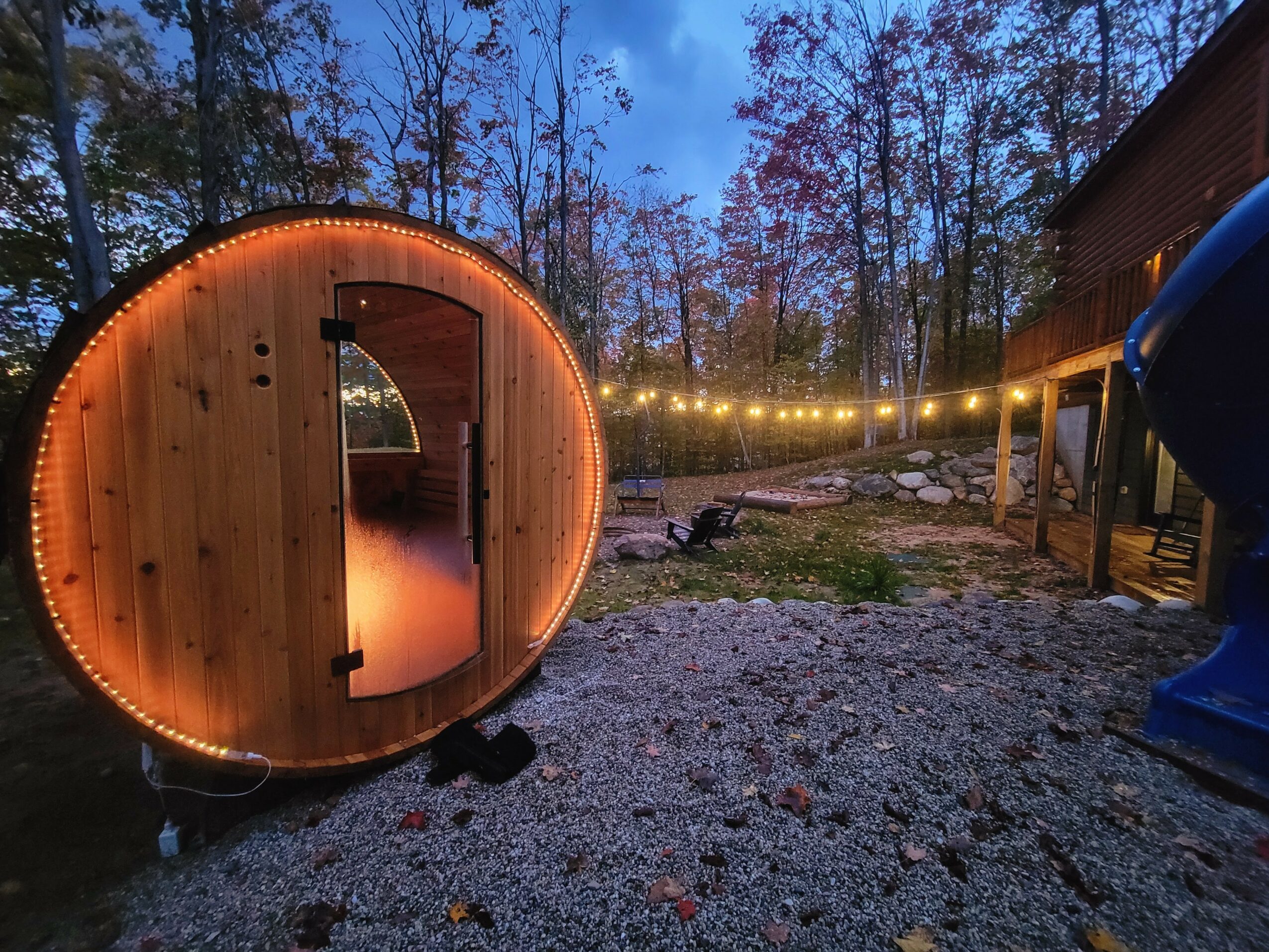 Wooden barrel-shaped sauna with glowing lights, outdoor setting with trees and string lights, gravel ground, evening sky.