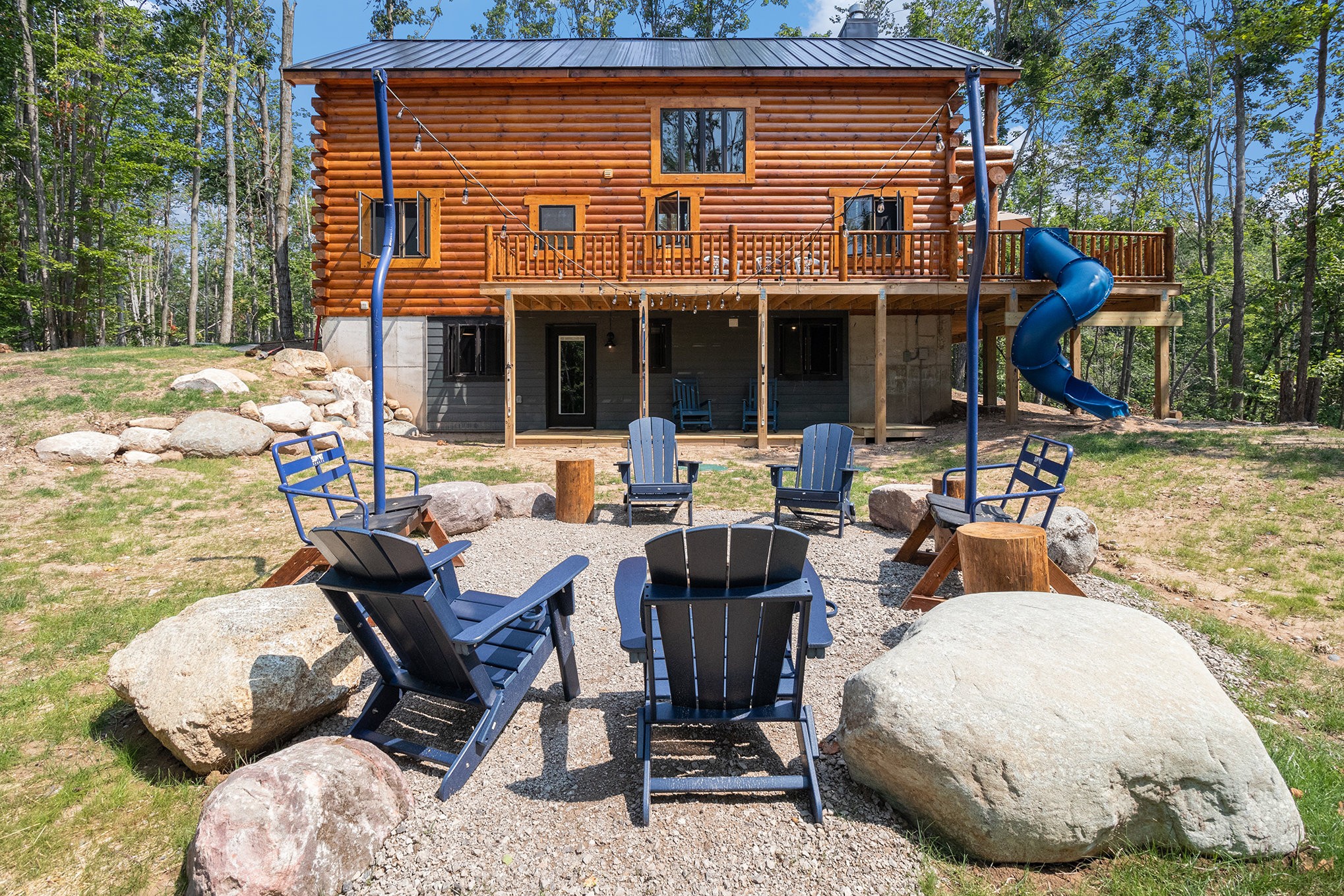 Backyard with a wooden house, a swing set, and a seating area with chairs and large rocks, surrounded by trees.