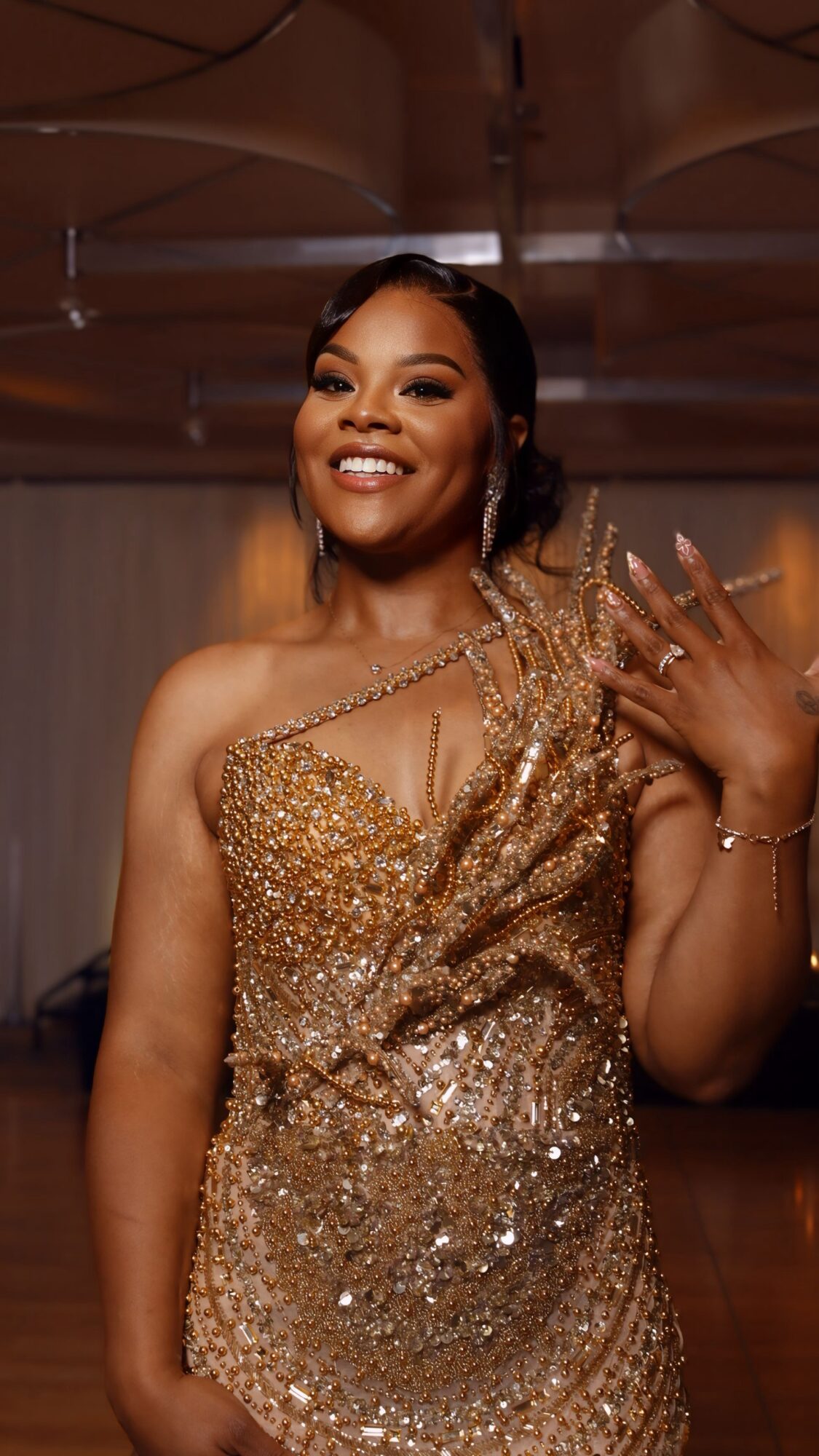 Smiling woman in a gold, sequined dress with one hand raised, in an indoor setting.