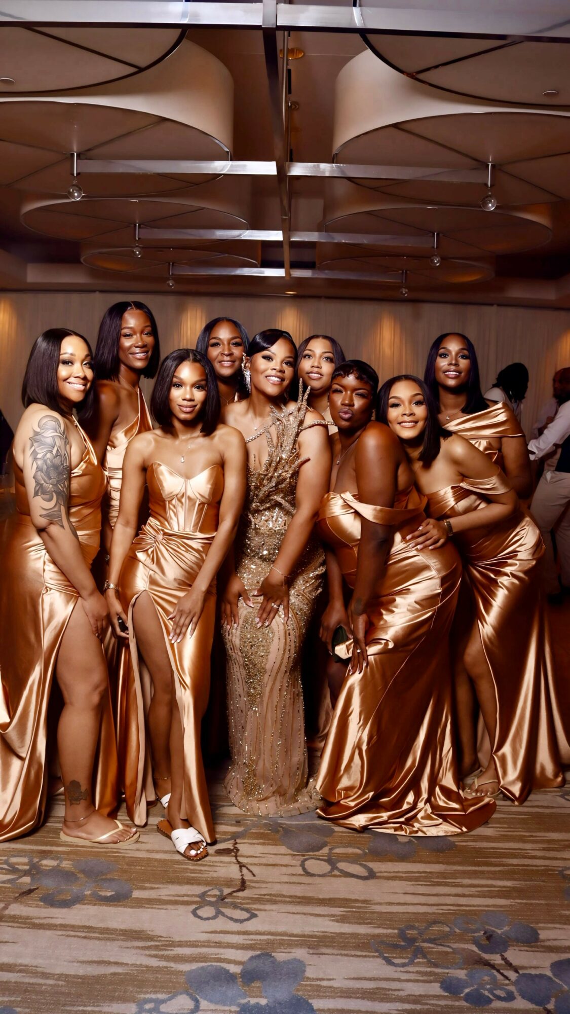 Group of women in matching bronze satin dresses posing indoors with ceiling design visible.
