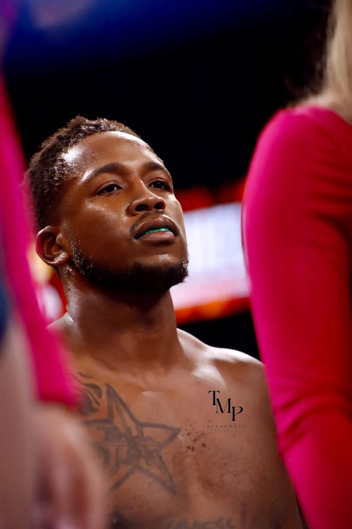 A shirtless man with tattoos looks upward, surrounded by people in pink clothing, in a dimly lit setting.