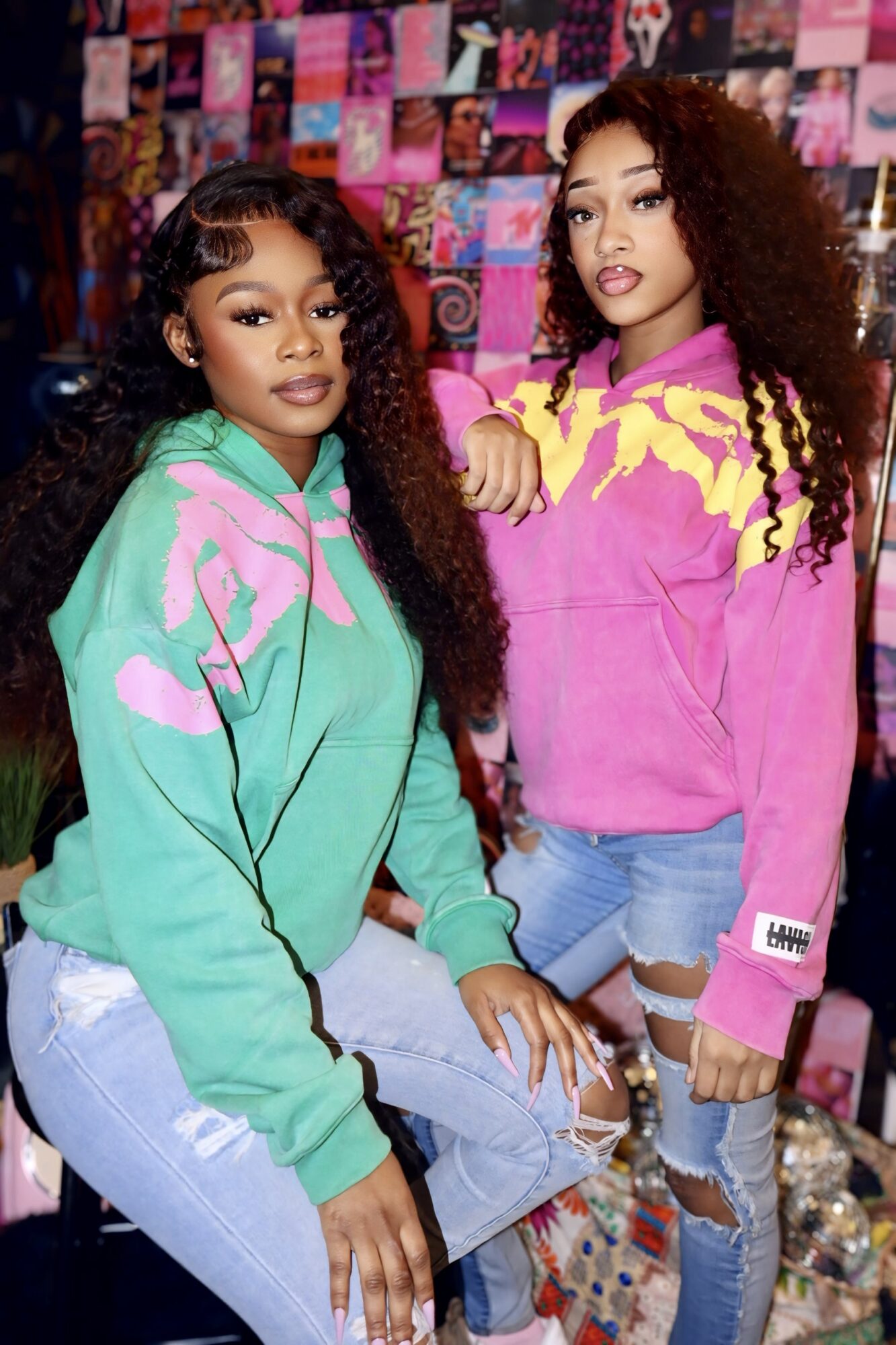 Two young women with curly hair pose in front of a colorful collage background, wearing vibrant hoodies and jeans.