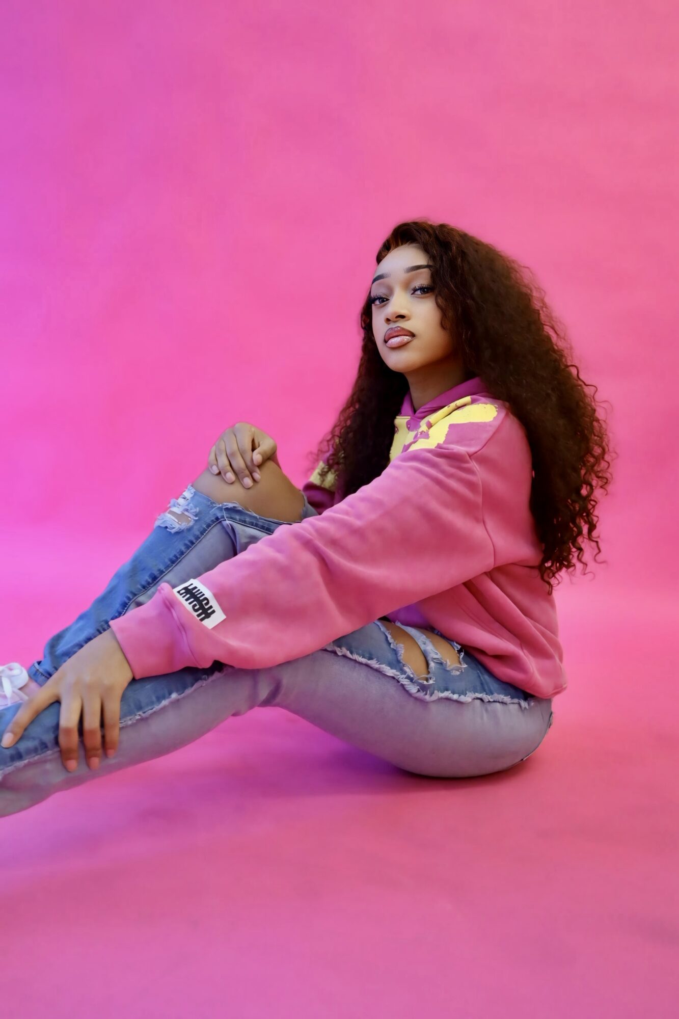 Young woman with curly hair sitting on pink background, wearing a pink jacket and ripped jeans, looking to the side.