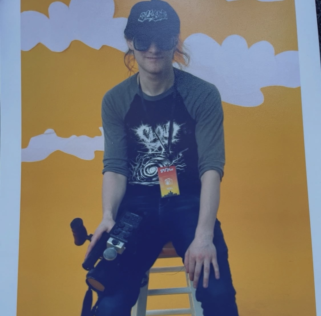 Person sitting on a stool with a camera, wearing sunglasses and a cap, against a yellow background with clouds.