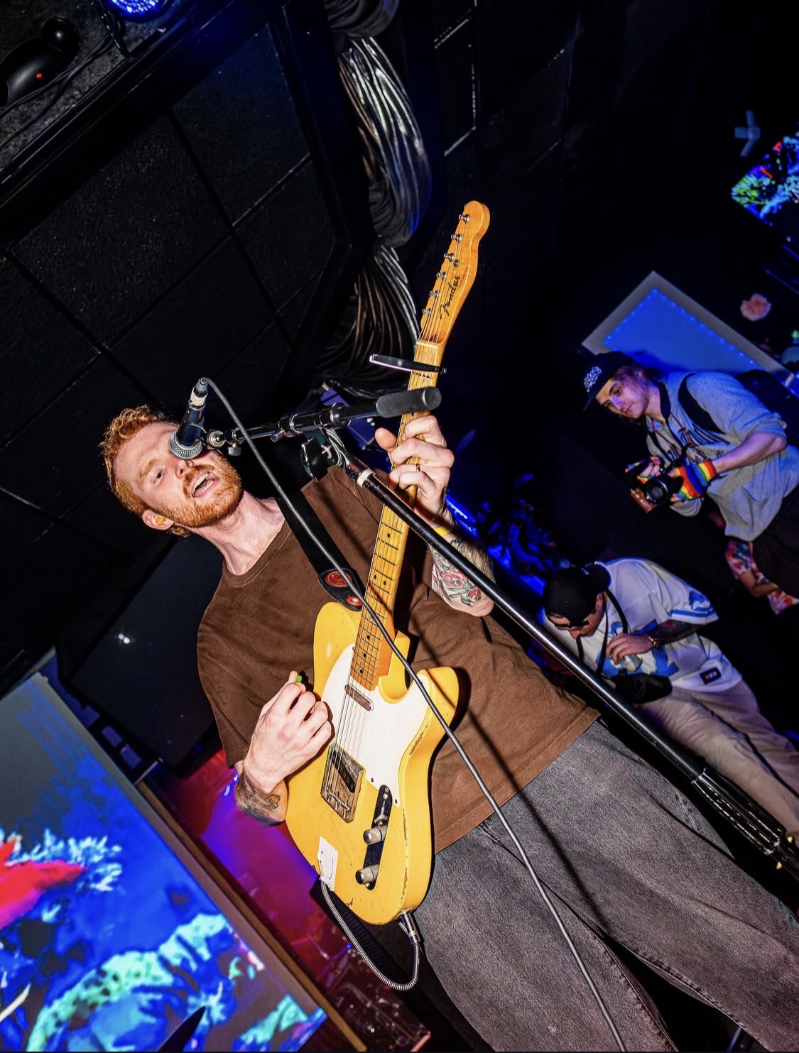 Person playing yellow electric guitar on stage, smiling, with microphone, audience in background, dim lighting, colorful screens.