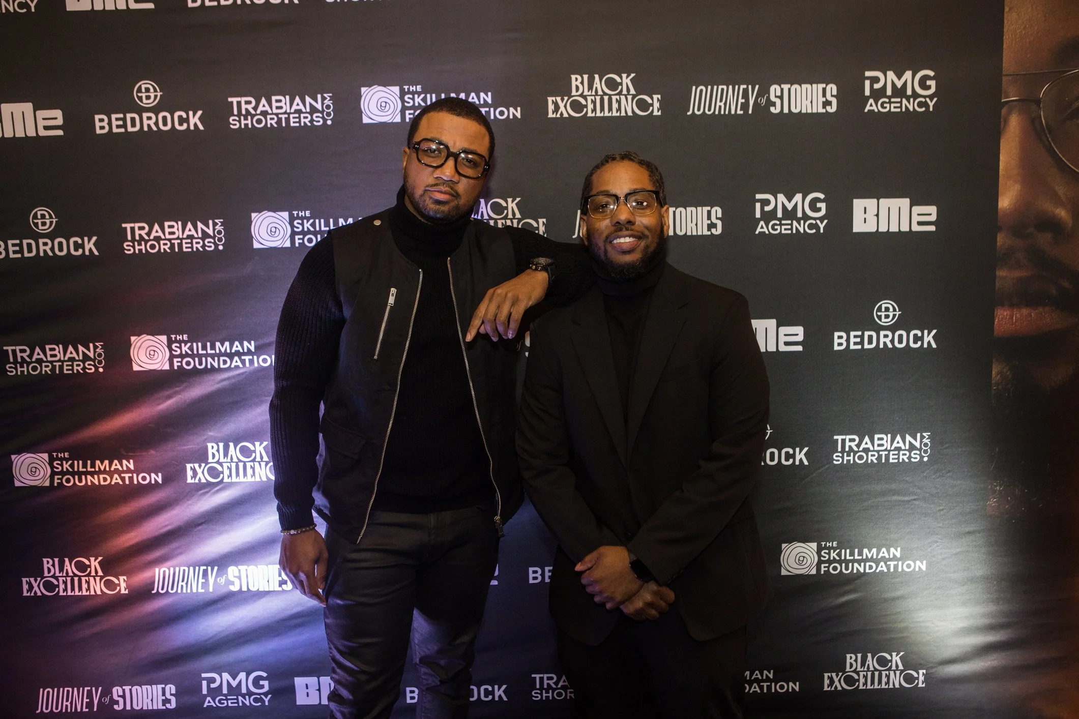 Two men standing in front of a black backdrop with logos, smiling, one with glasses and beard, wearing a black jacket.