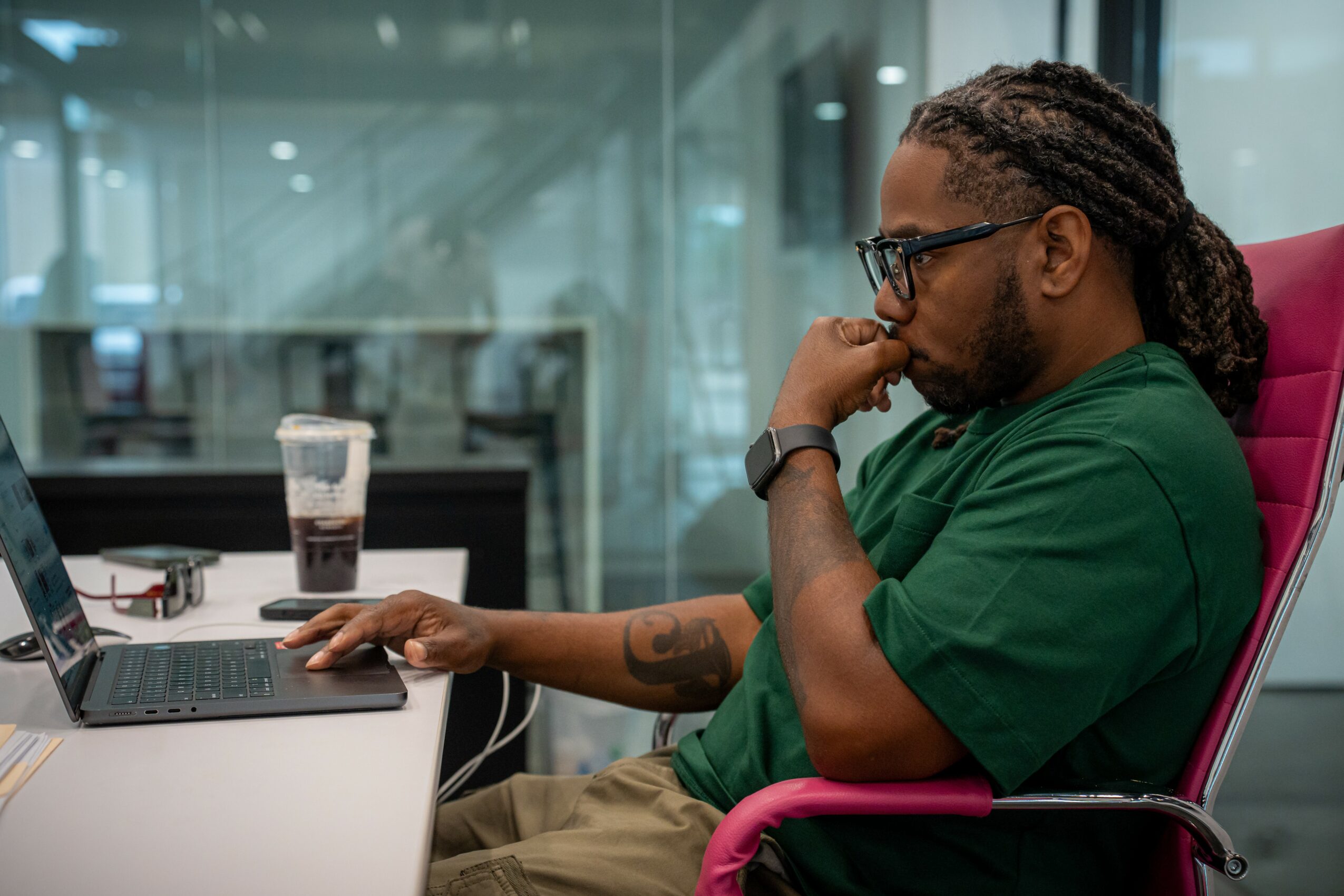Man with glasses and dreadlocks working on a laptop in an office, with a drink on the desk.