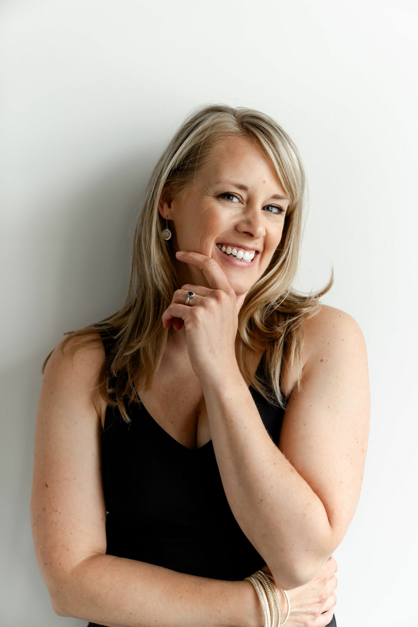 Smiling woman with blonde hair wearing a black top, touching her chin, against a plain white background.