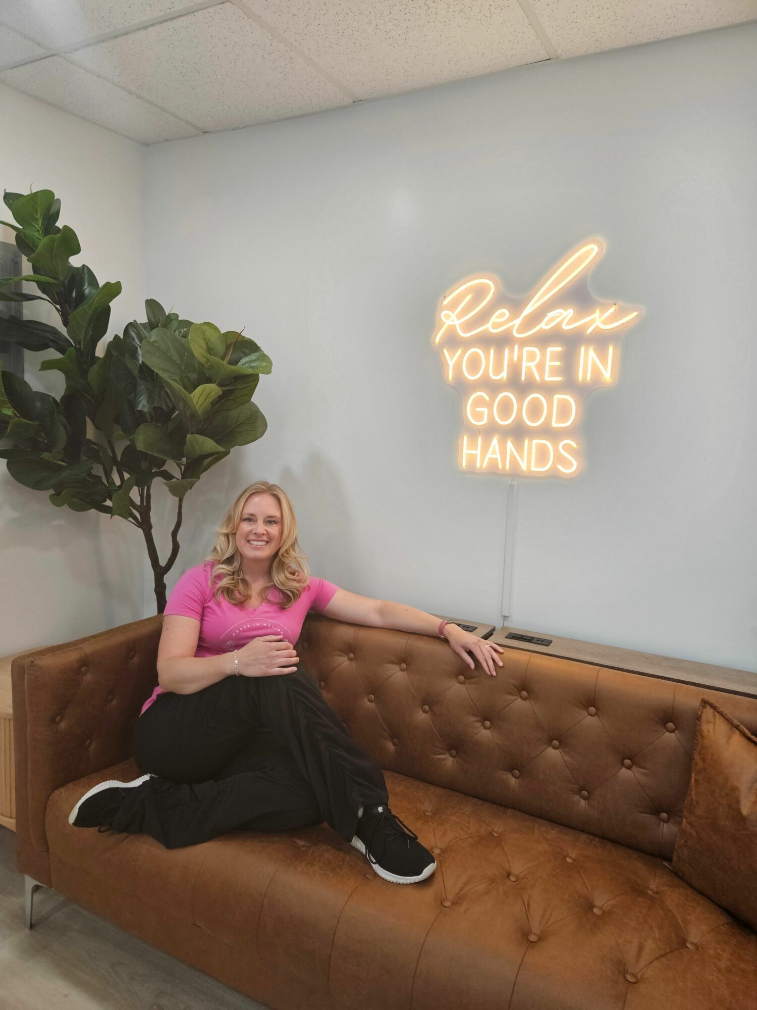Woman sitting on a brown leather couch with a plant nearby, smiling, in front of a wall with neon sign reading 'Relax, you're in good hands'.