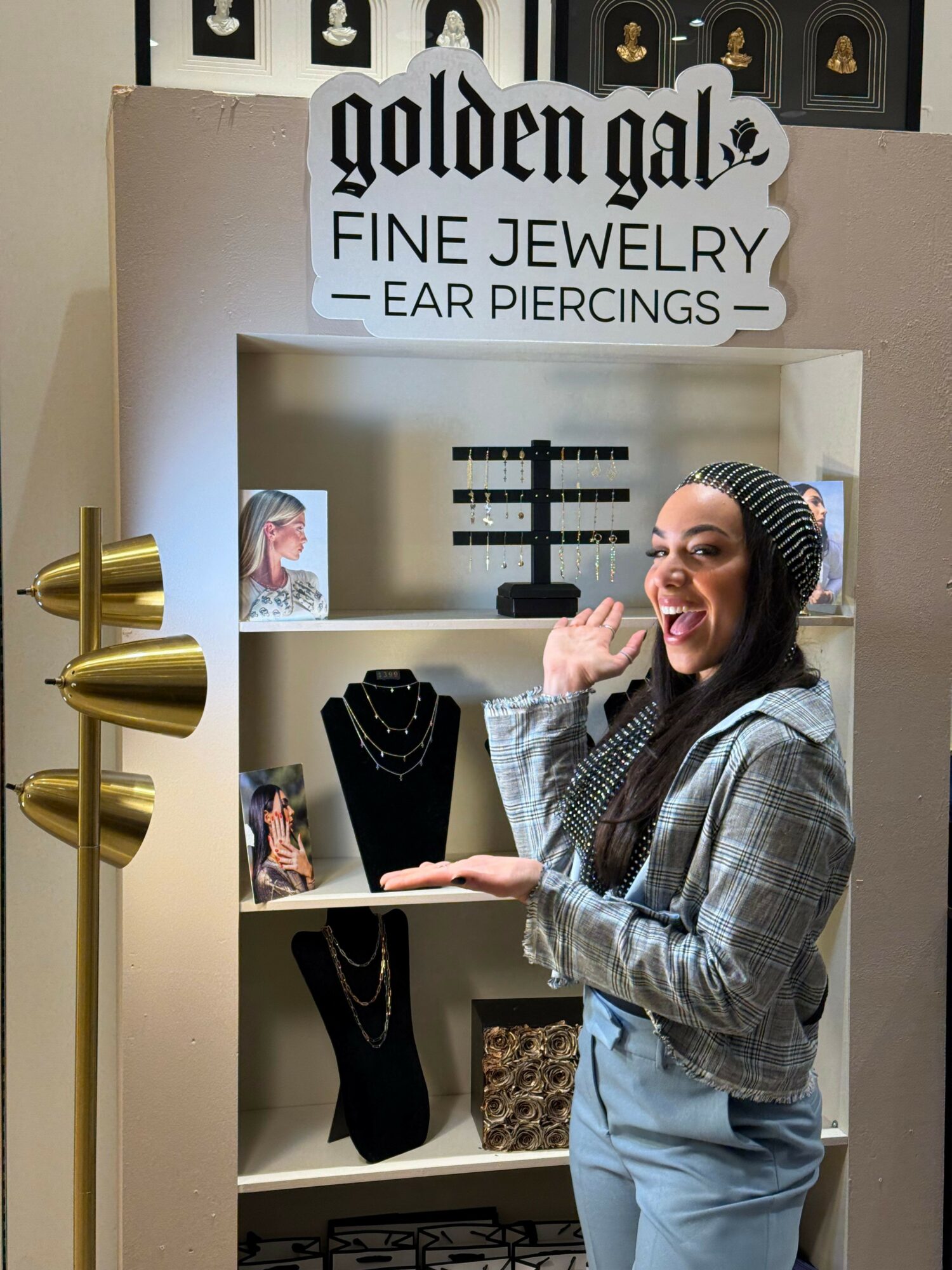 Person with long hair and striped hat gestures near jewelry display in store. Sign reads 'Gotham Fine Jewelry & Ear Piercings.'