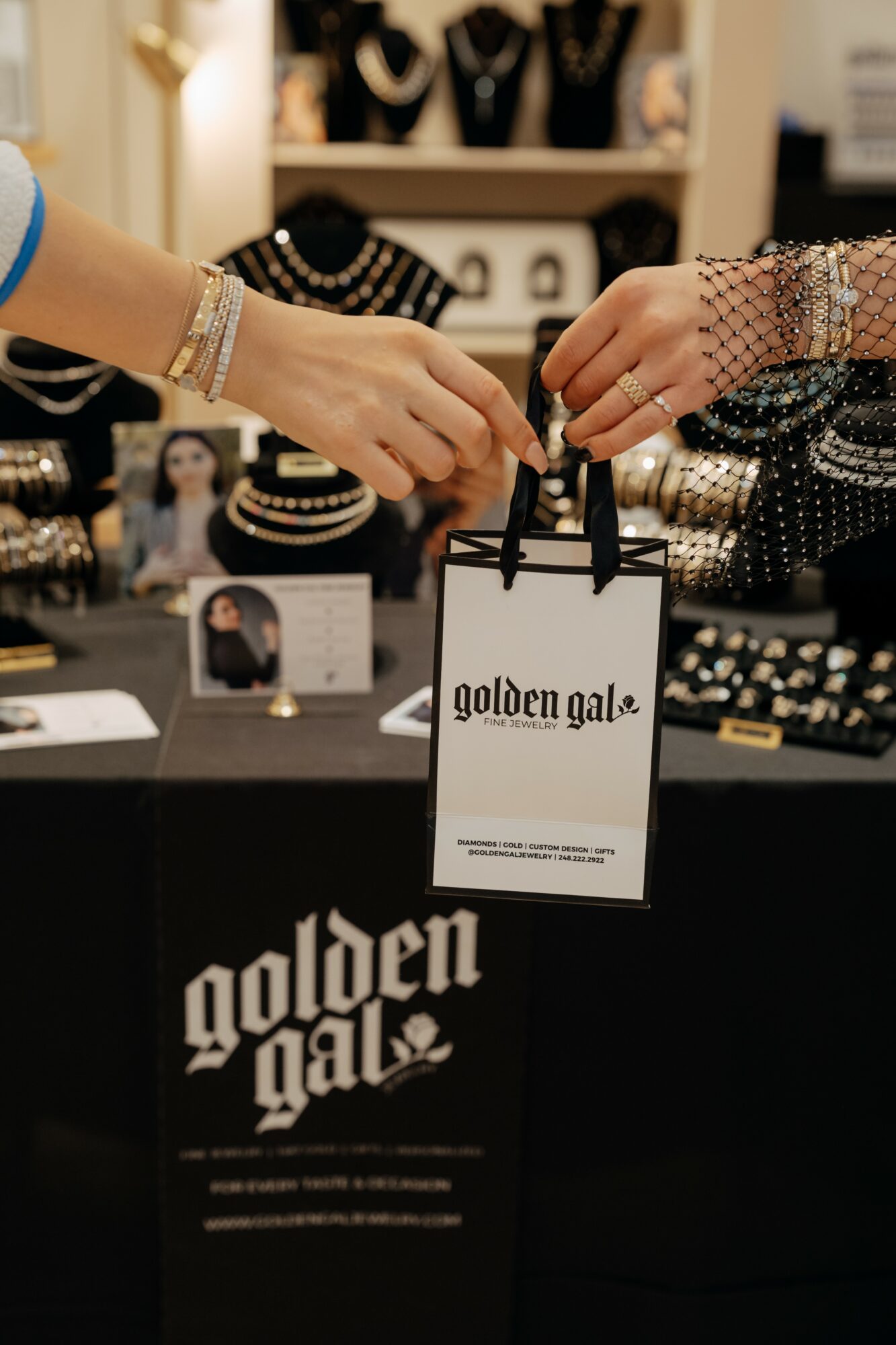 Two people exchange a shopping bag at a jewelry display, with jewelry and accessories in the background.
