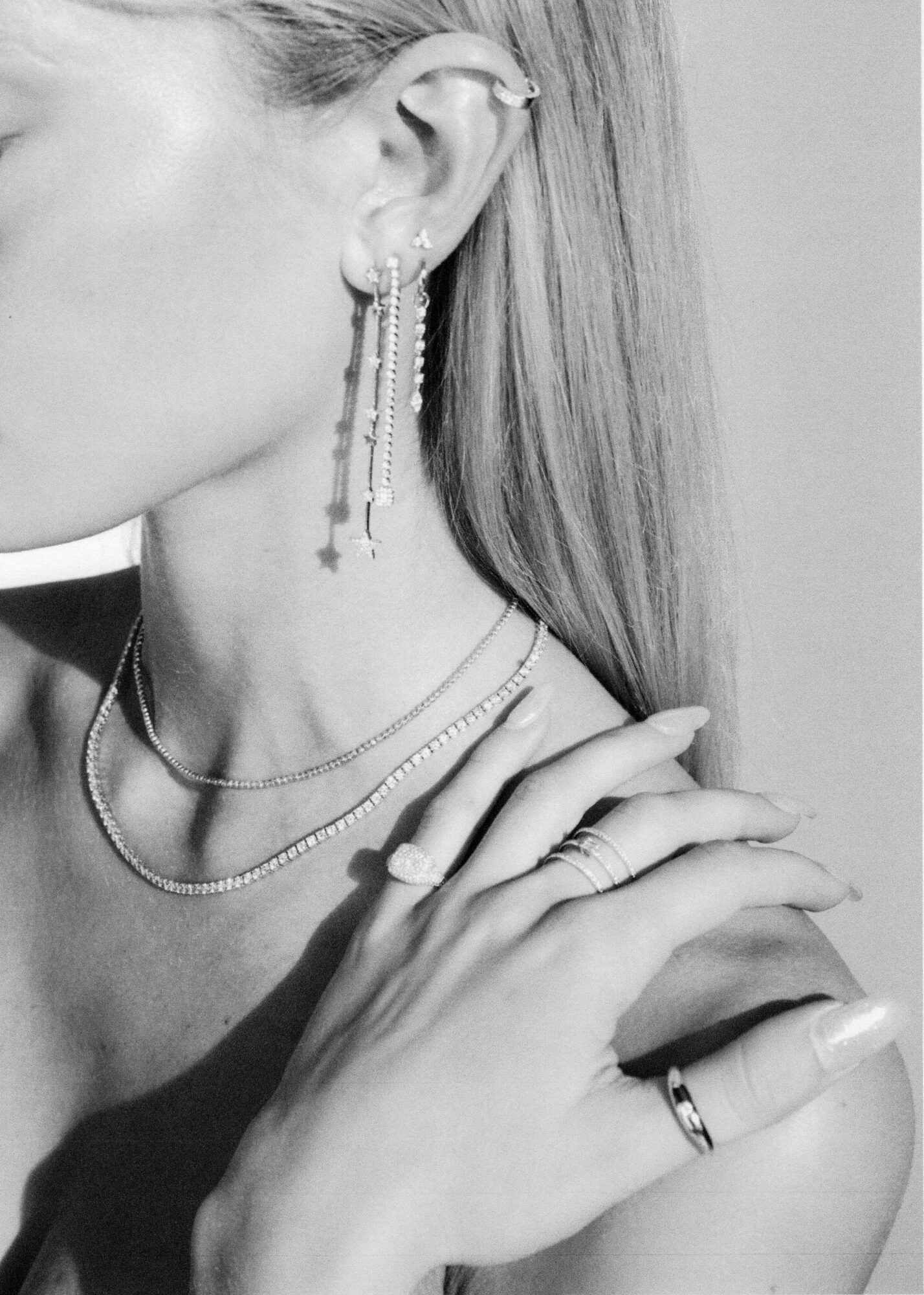 Close-up of a woman's ear with dangling earrings, layered necklaces, and rings on her fingers, in black and white.