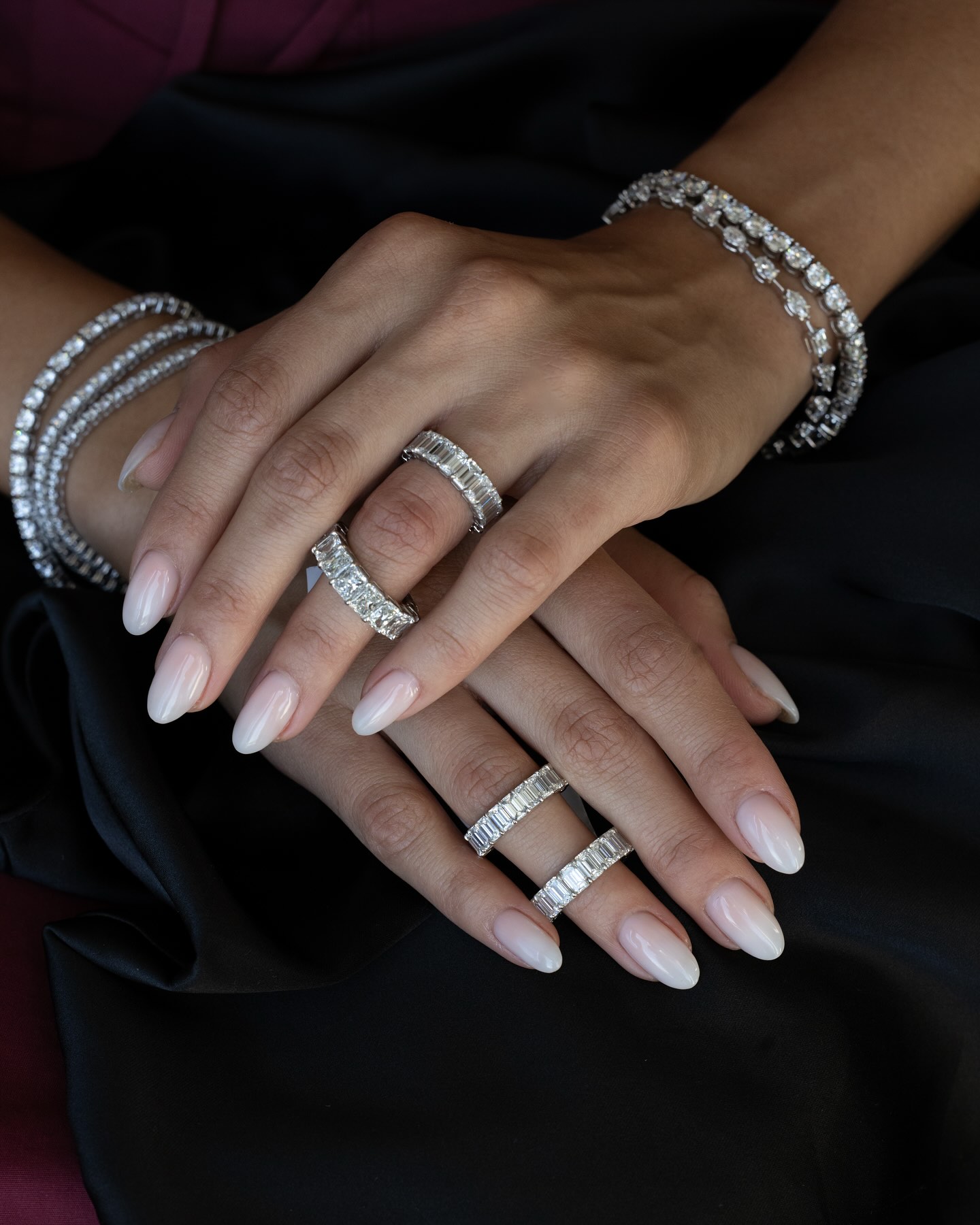 Two hands with multiple diamond rings and bracelets resting on a black fabric surface, nails painted light pink.