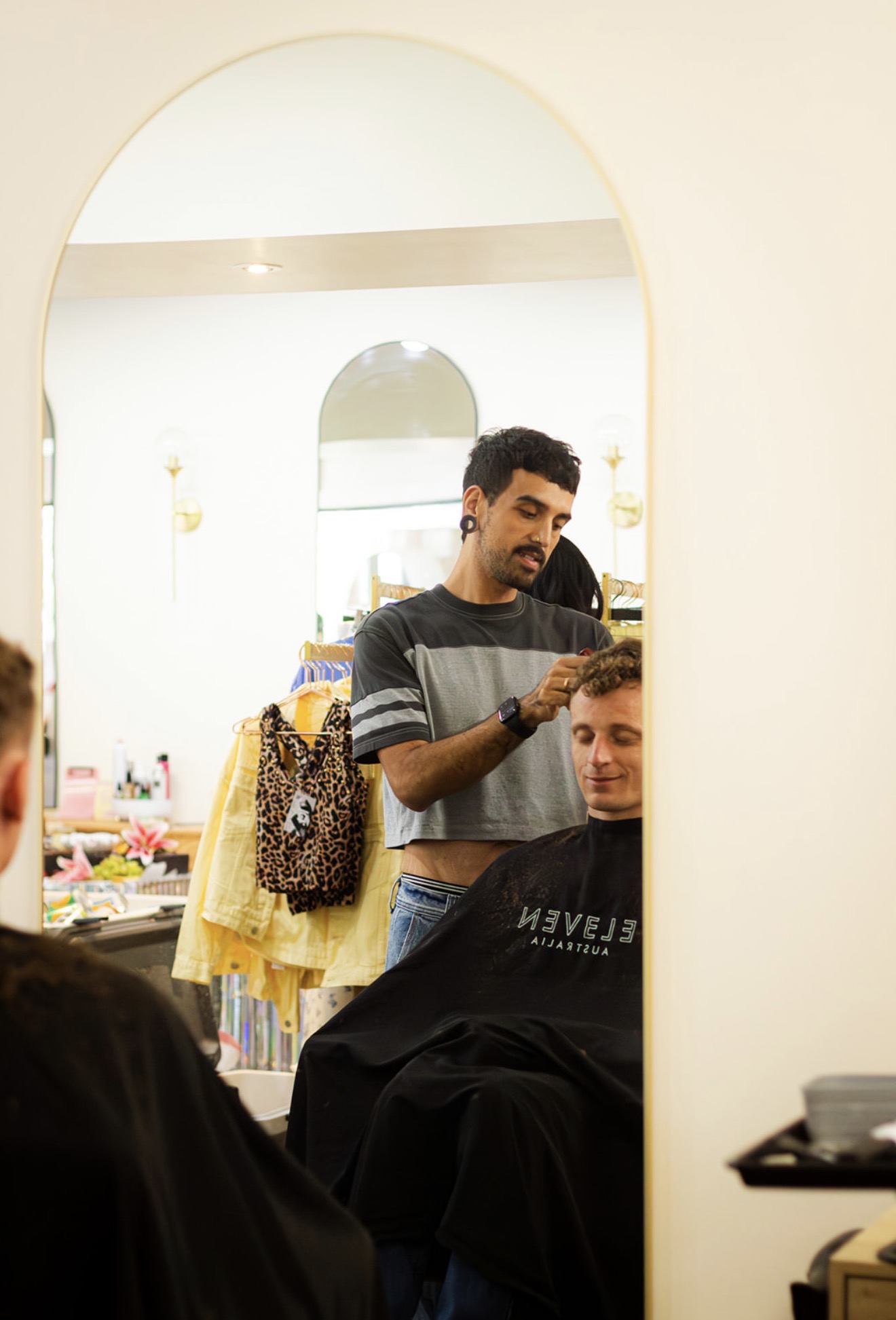 Person getting a haircut in a salon, stylist working on client's hair, mirror reflecting the scene, clothing and salon items visible.
