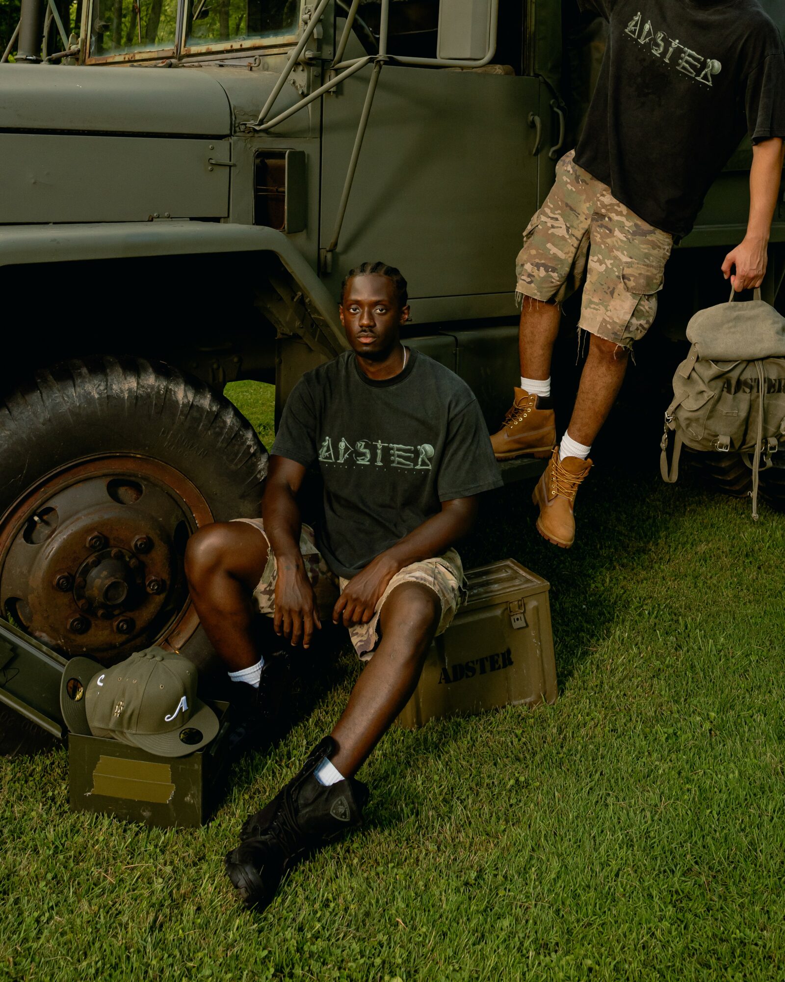 Person sitting on a box next to a military vehicle, wearing a black shirt, shorts, and black boots, with another person standing nearby.