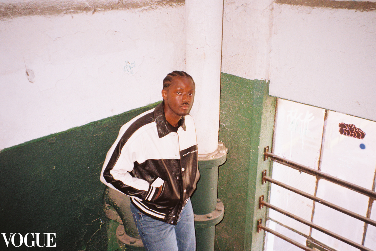 Person with braided hair wearing a black and white jacket and jeans, standing in a stairwell with green and white walls.