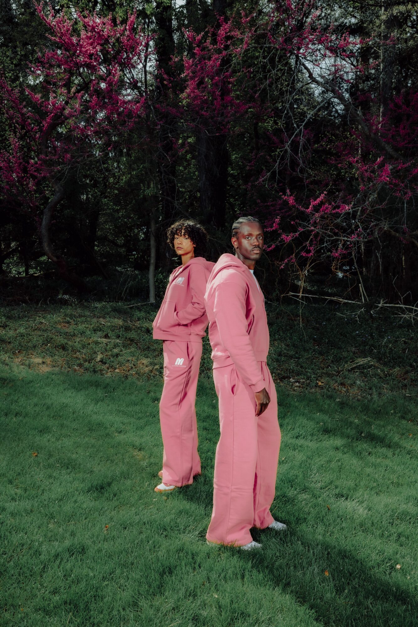 Two people in pink outfits standing on grass with trees and pink blossoms in background.