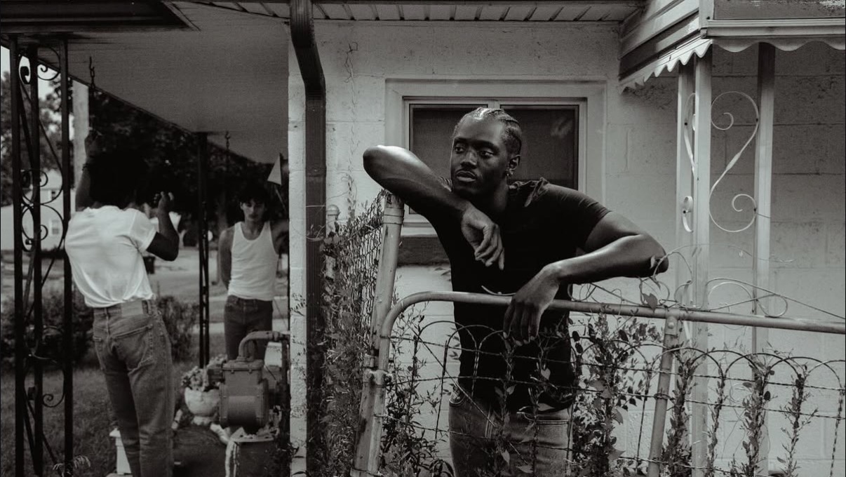 Man leaning on a fence outside a house, with two people in the background near a porch, black and white photo.