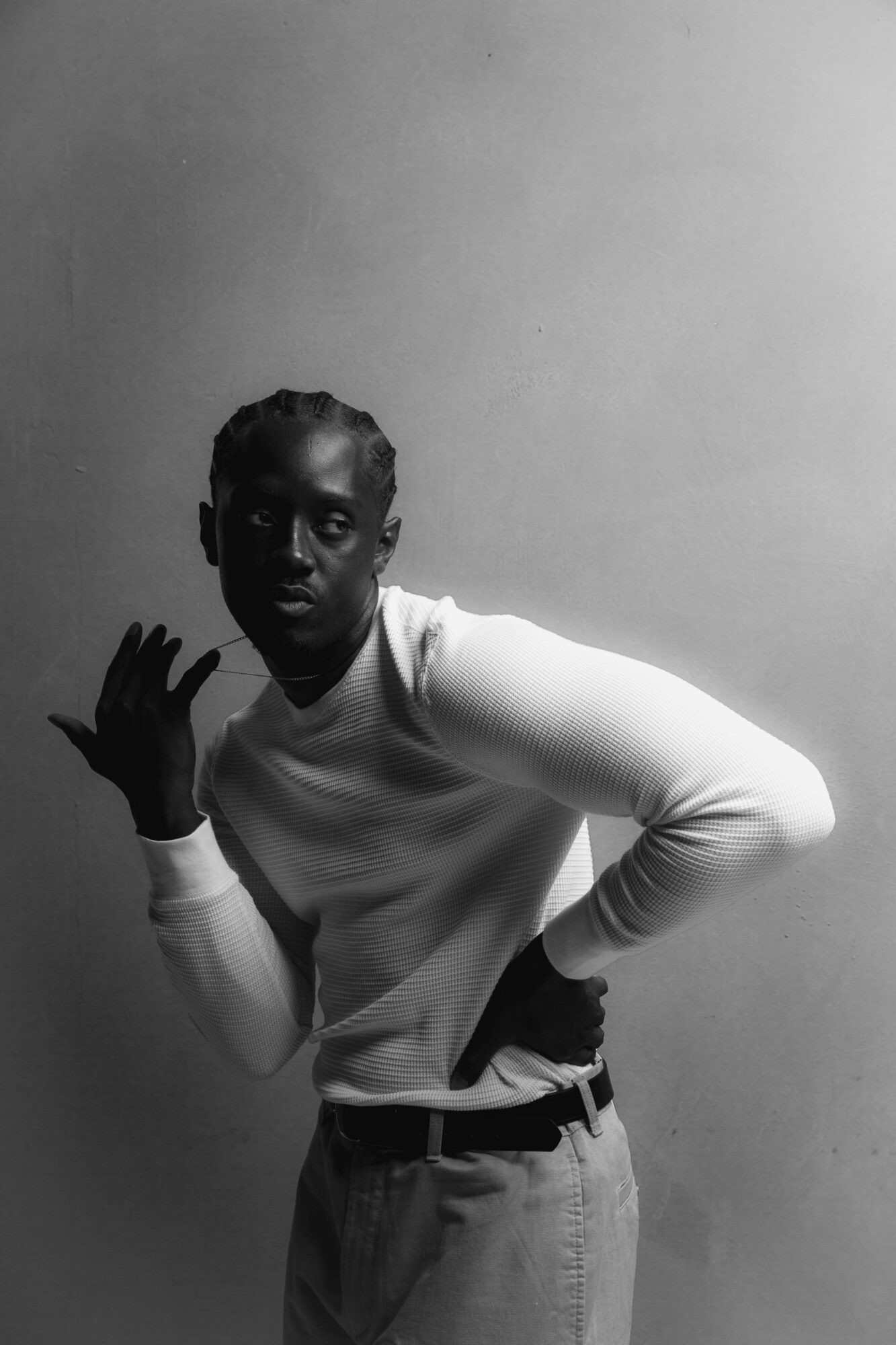 Person with braided hair wearing a white long-sleeve shirt and pants, standing against a plain wall.