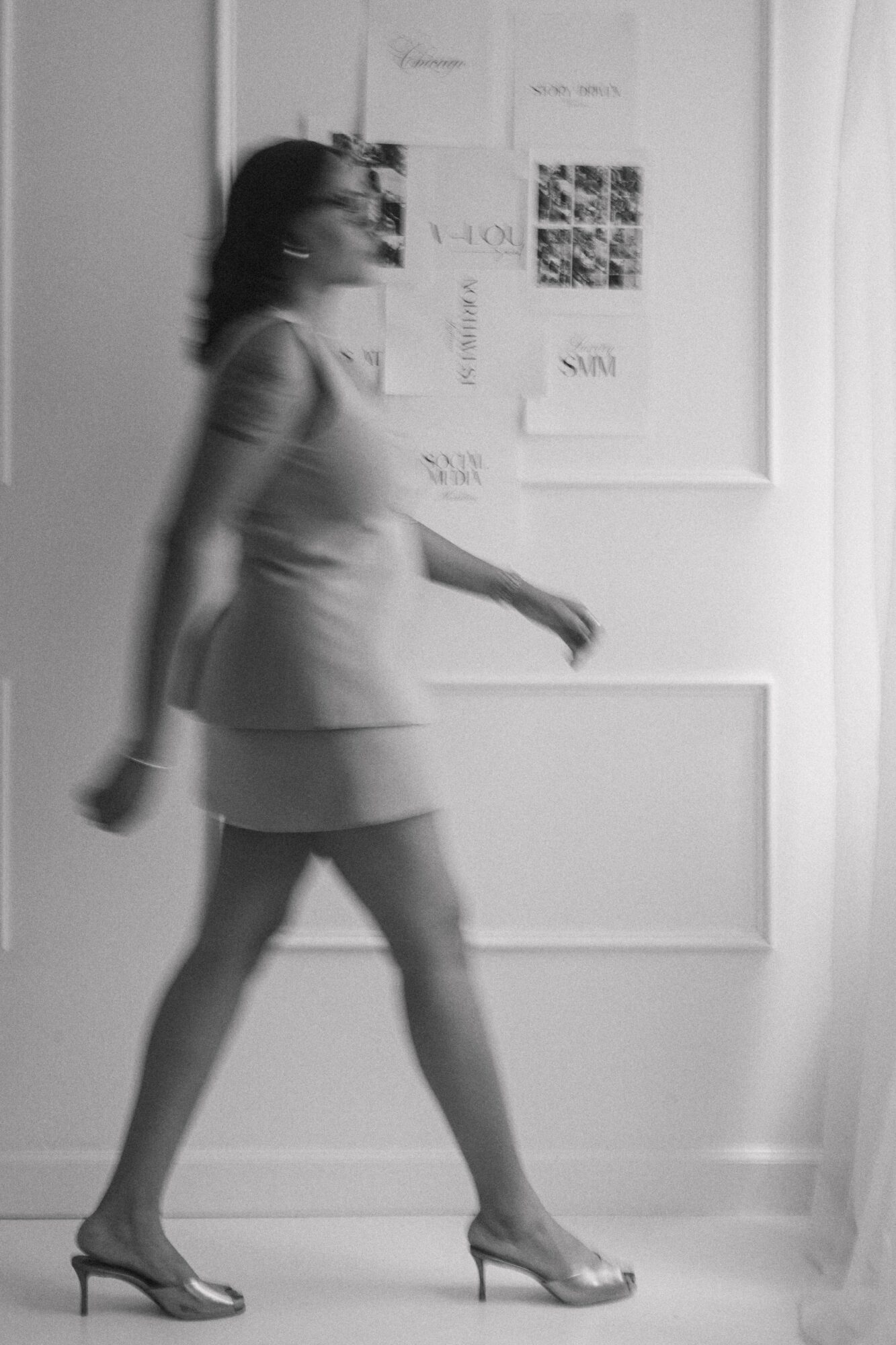 Blurry woman walking in front of a wall with papers and images, in a room with white walls and curtains.