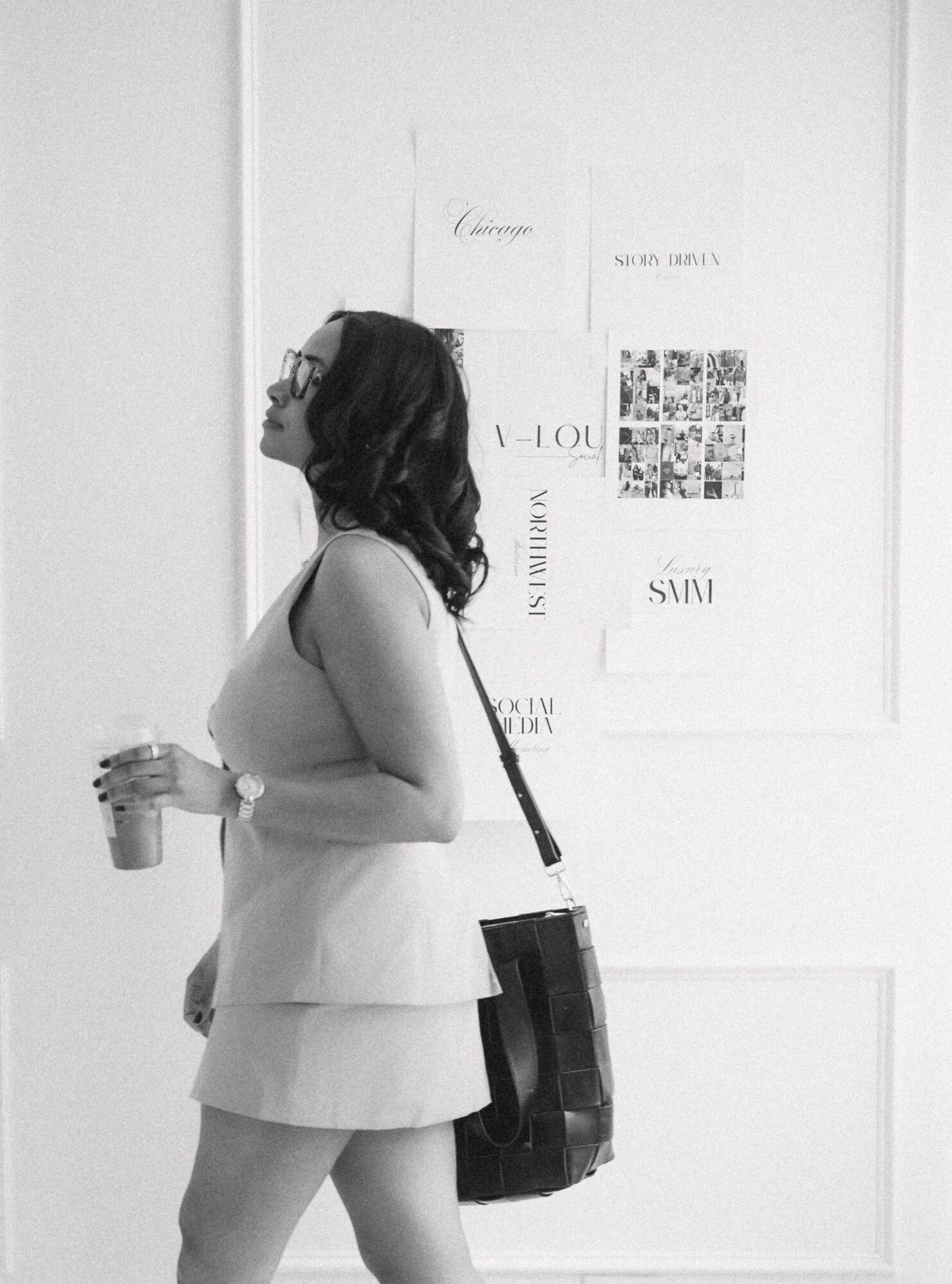 Woman with glasses holding a drink, standing in front of a wall with framed artwork, wearing a layered dress and carrying a bag.