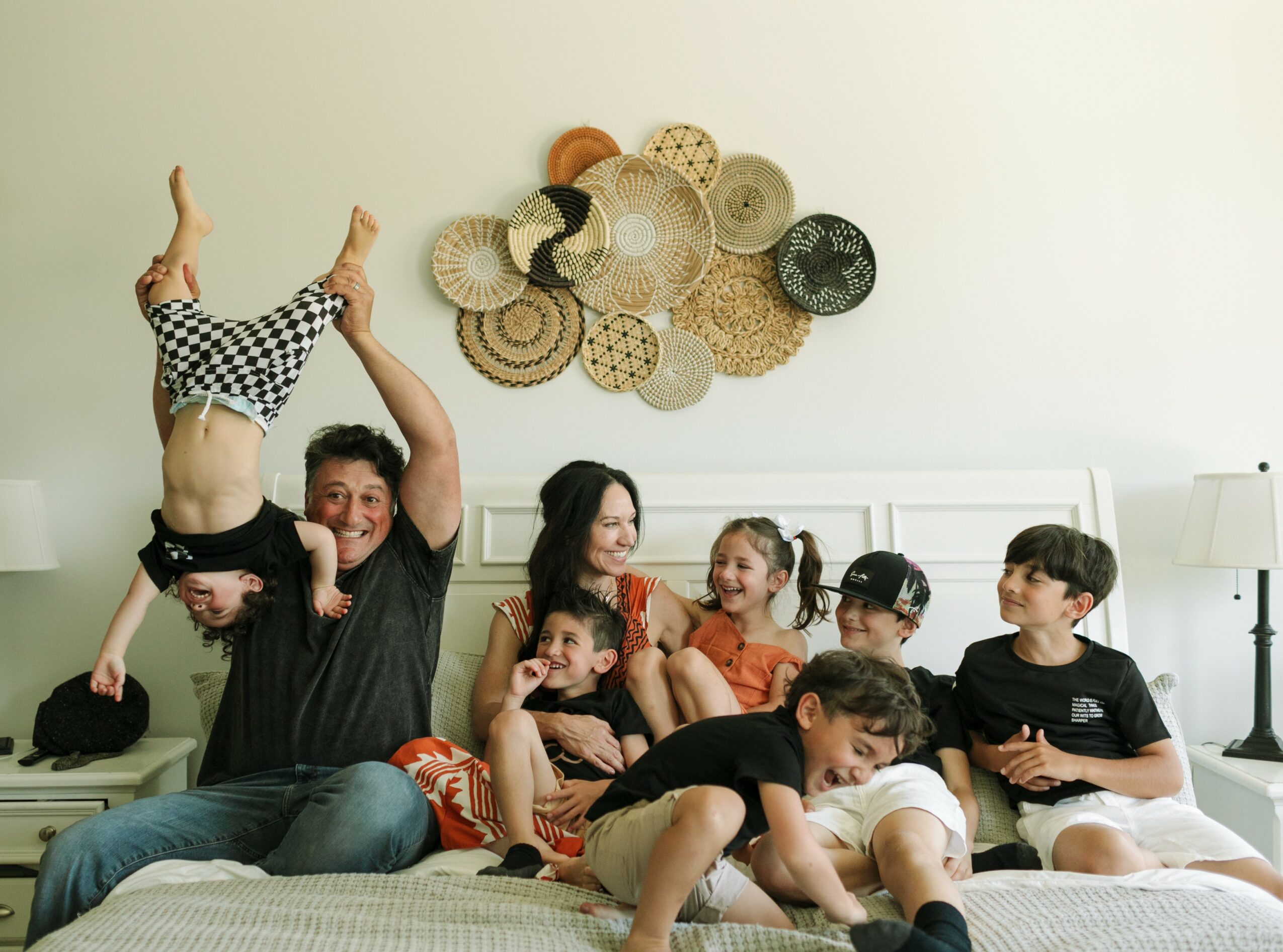 Group of children and adults on a bed, with one person holding a child upside down, smiling and playing, wall decor above.