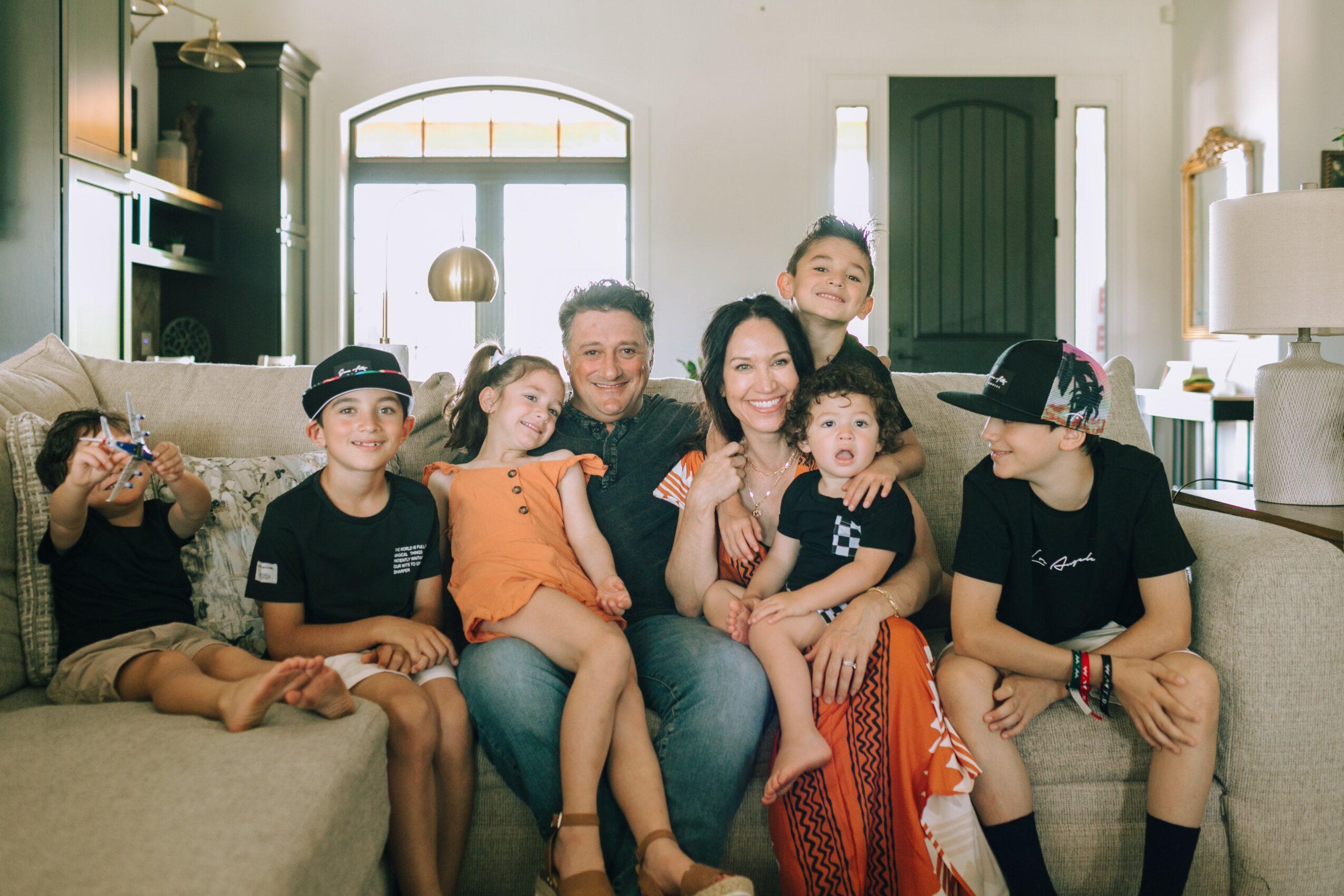 Family of seven sitting on a sofa in a living room, smiling and posing for the photo.