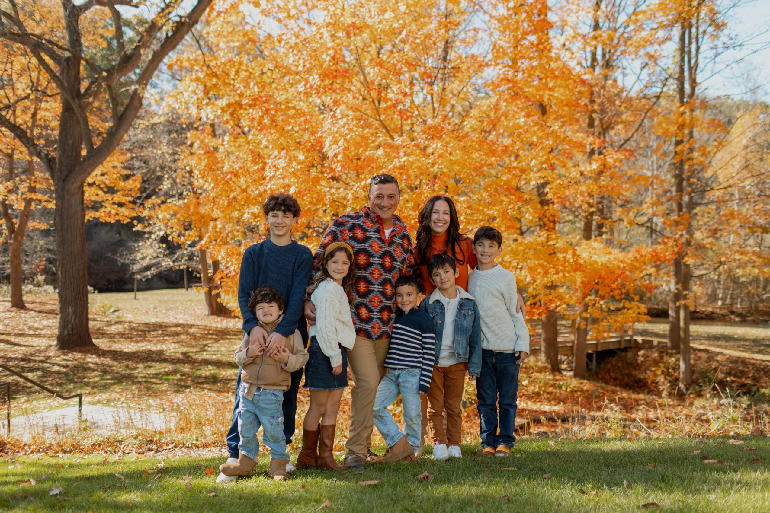 Group of seven people standing outdoors in a park with autumn trees and fallen leaves, smiling and posing for a photo.