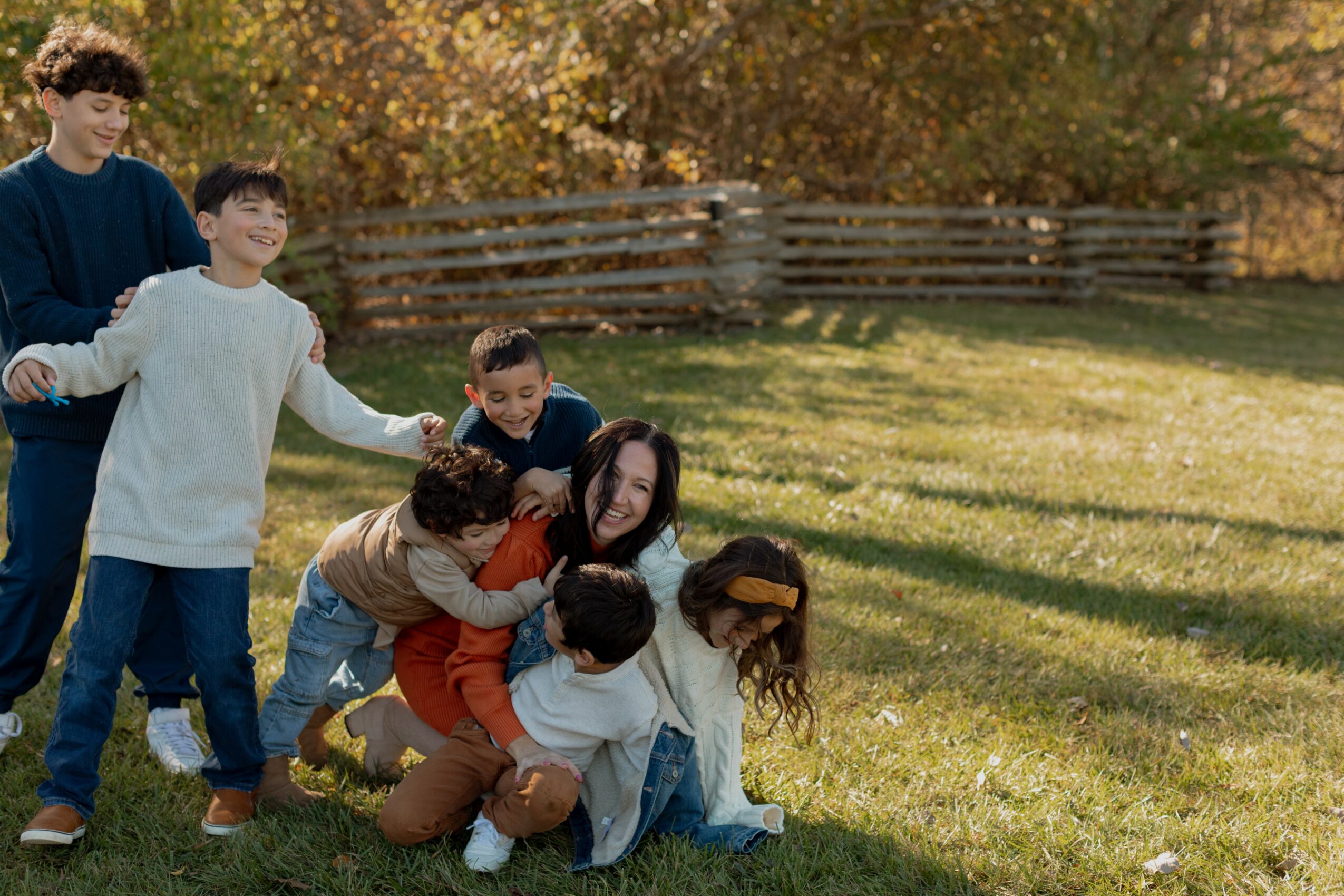 Group of children and a woman playing outdoors on grass with trees and a fence in background.
