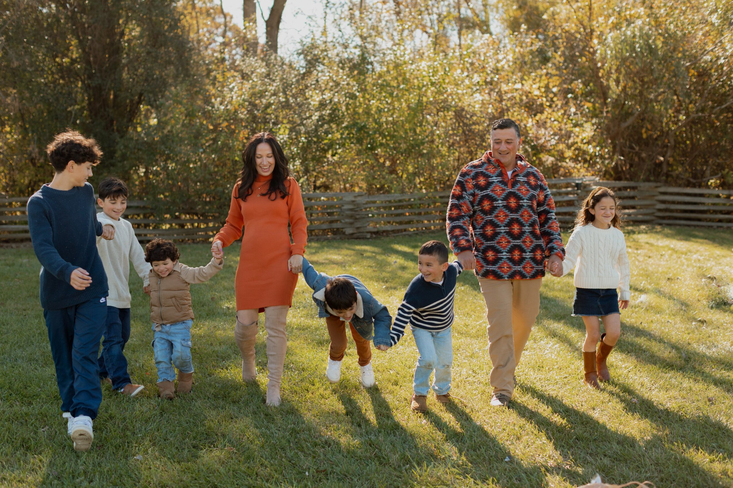 Group of children and two adults holding hands and playing outdoors in a grassy area with trees.