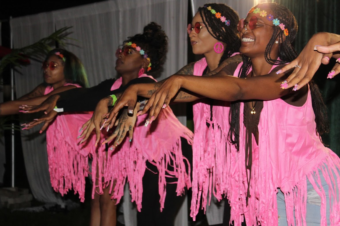Group of women in pink fringed dresses with colorful headbands, dancing with arms extended, smiling, indoors.