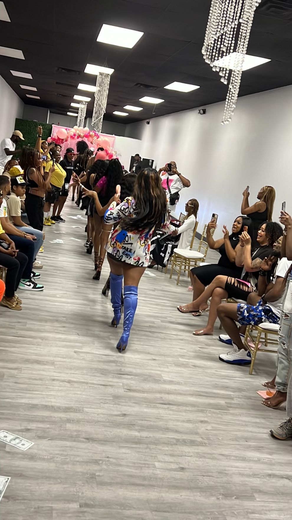 People sitting and standing in a room with a chandelier, some using phones, others talking, with a wooden floor.