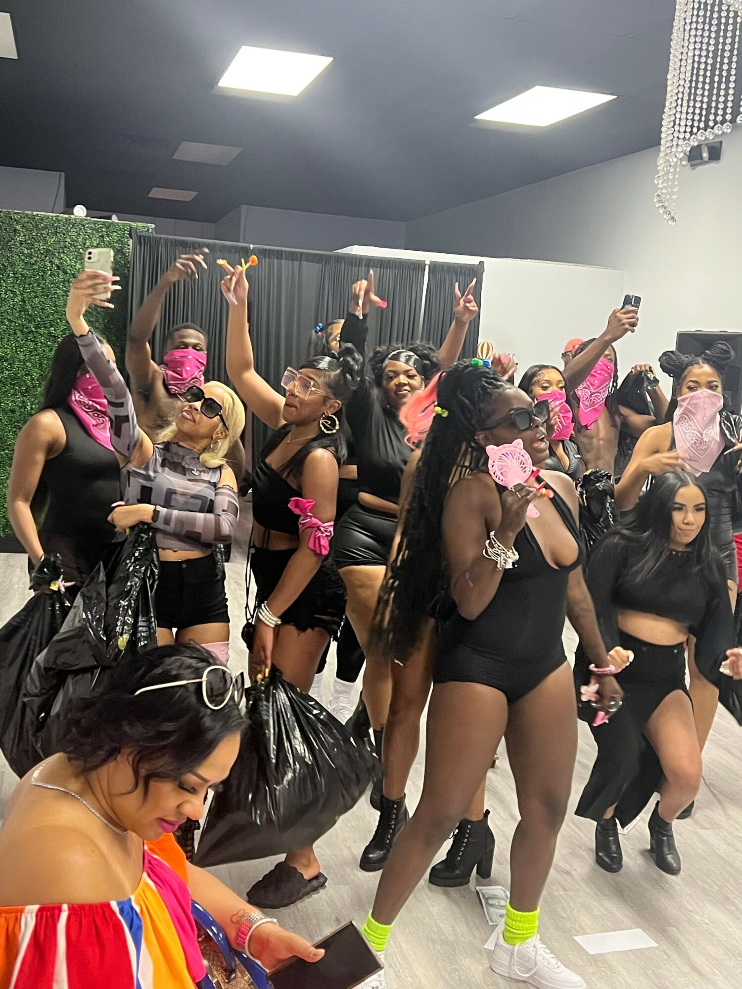 Group of women in black outfits and colorful accessories posing in a room with black curtains and ceiling lights.