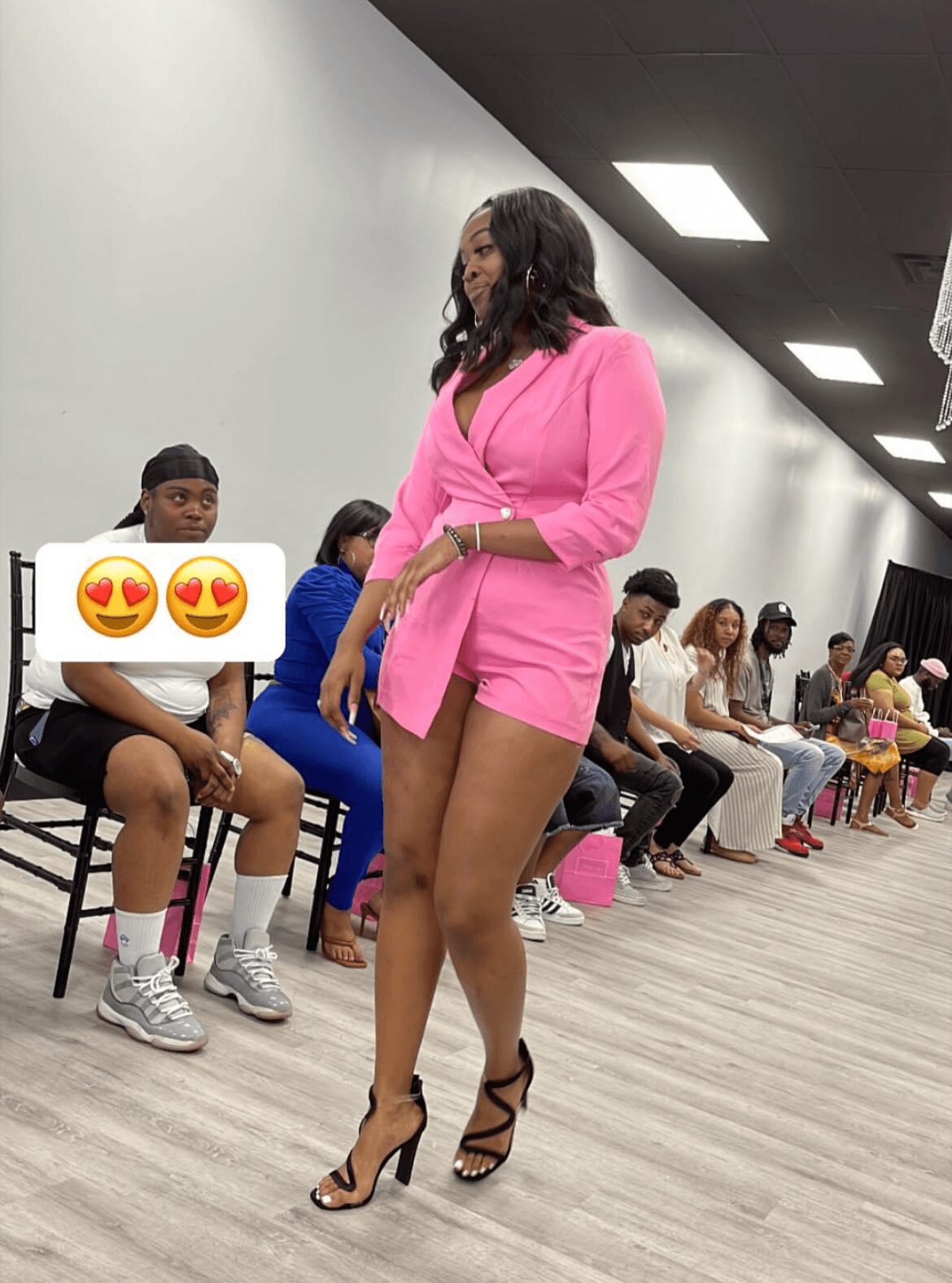 Woman in pink blazer and shorts standing in a room with seated people, some holding phones, in a line along the wall.