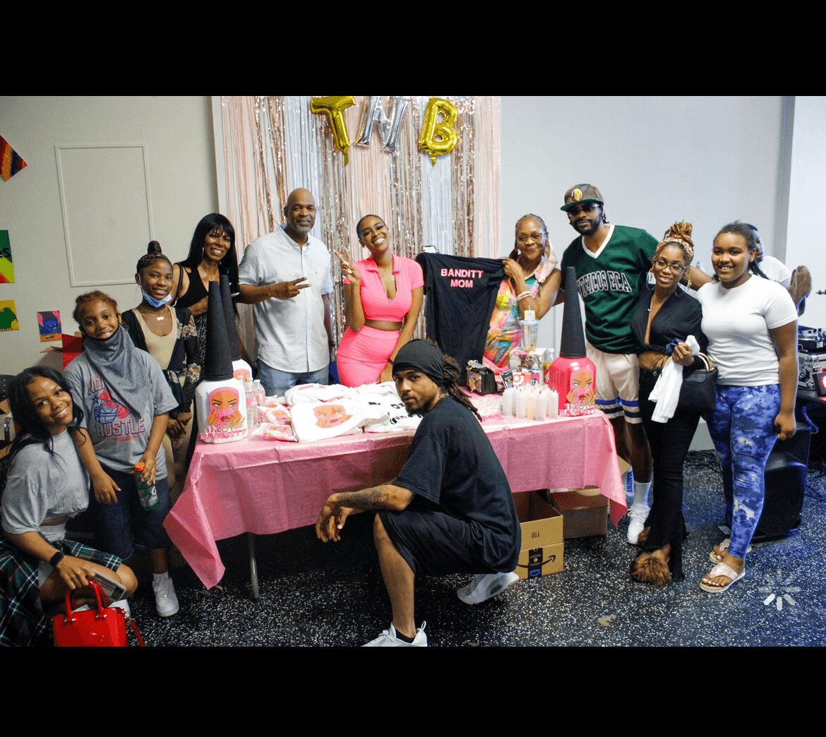 Group of people celebrating around a table with pink tablecloth, balloons, and gifts, in a decorated room.