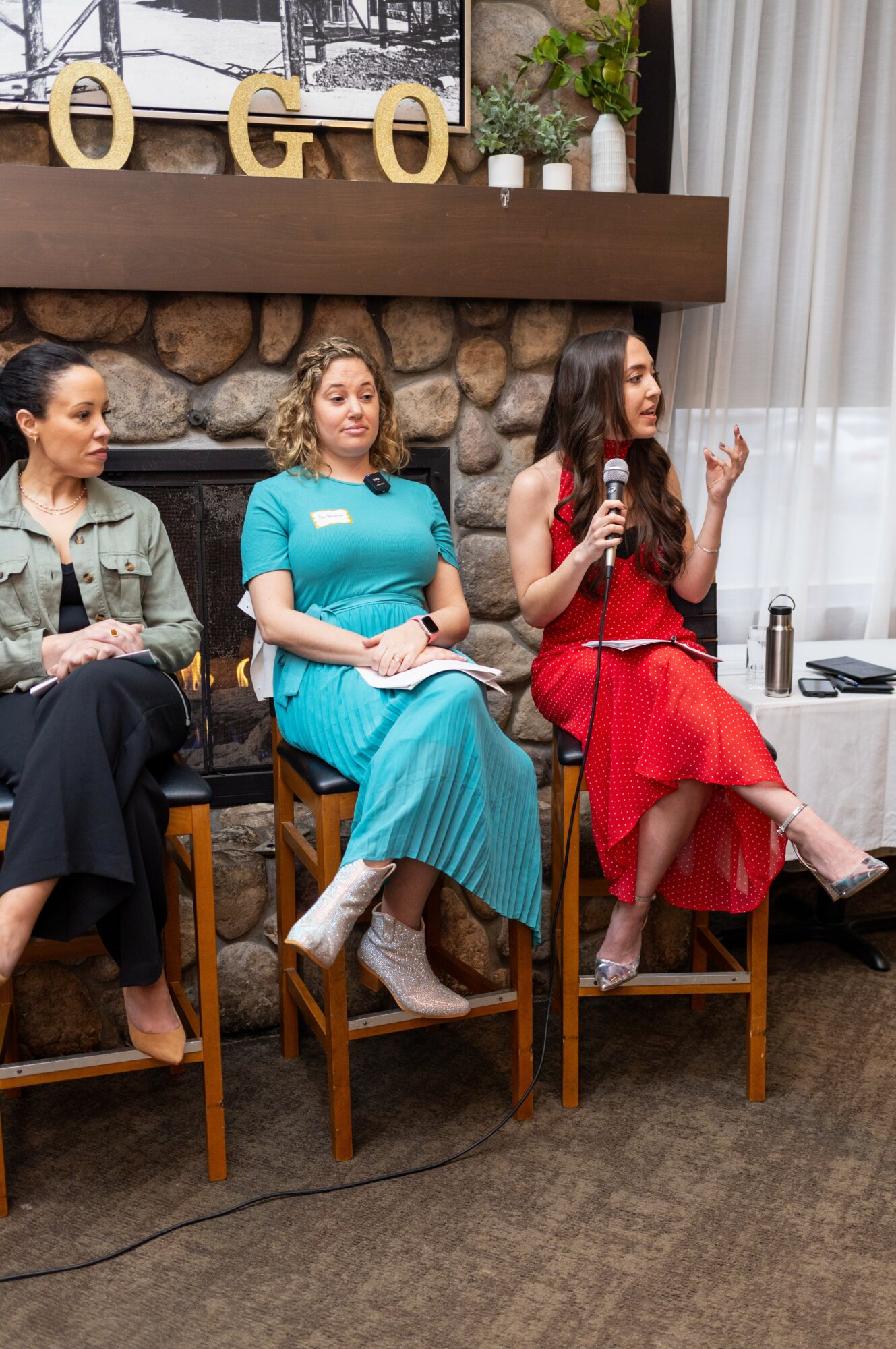 Three women seated on chairs, one speaking into a microphone, in a room with a stone fireplace and curtains.