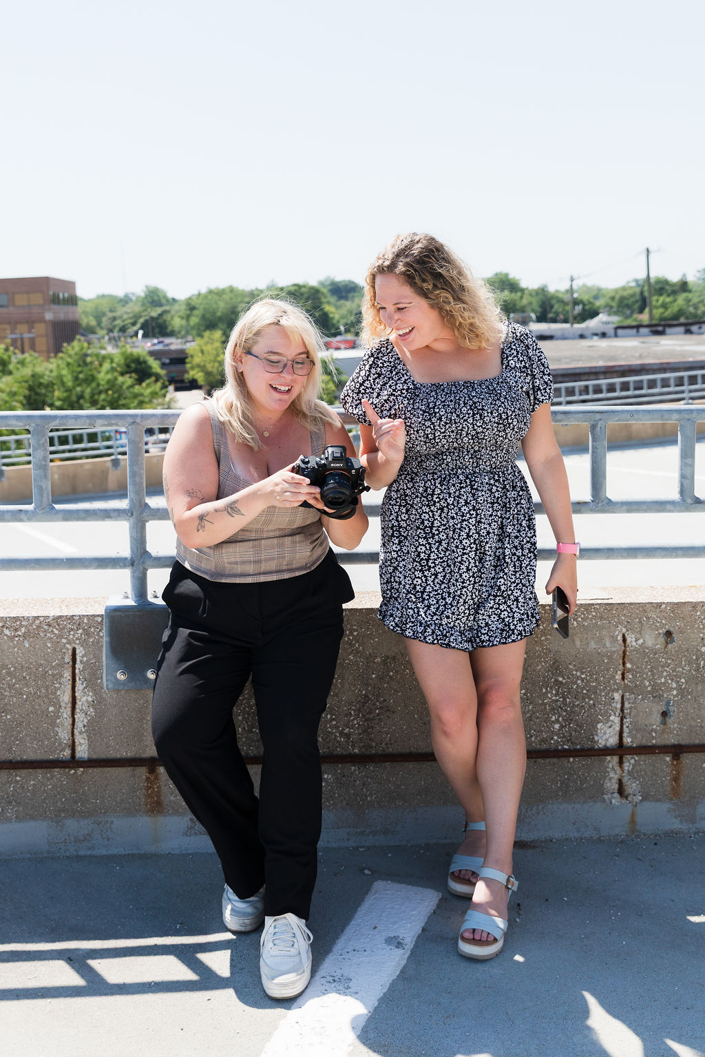 Two women stand outdoors on a sunny day, looking at a camera, with a railing and cityscape in the background.