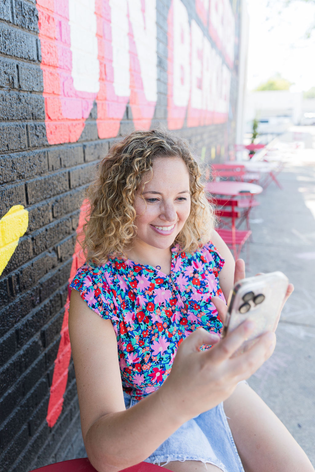 Young woman with curly hair smiling while looking at her phone, sitting outdoors against a brick wall with pink lettering.