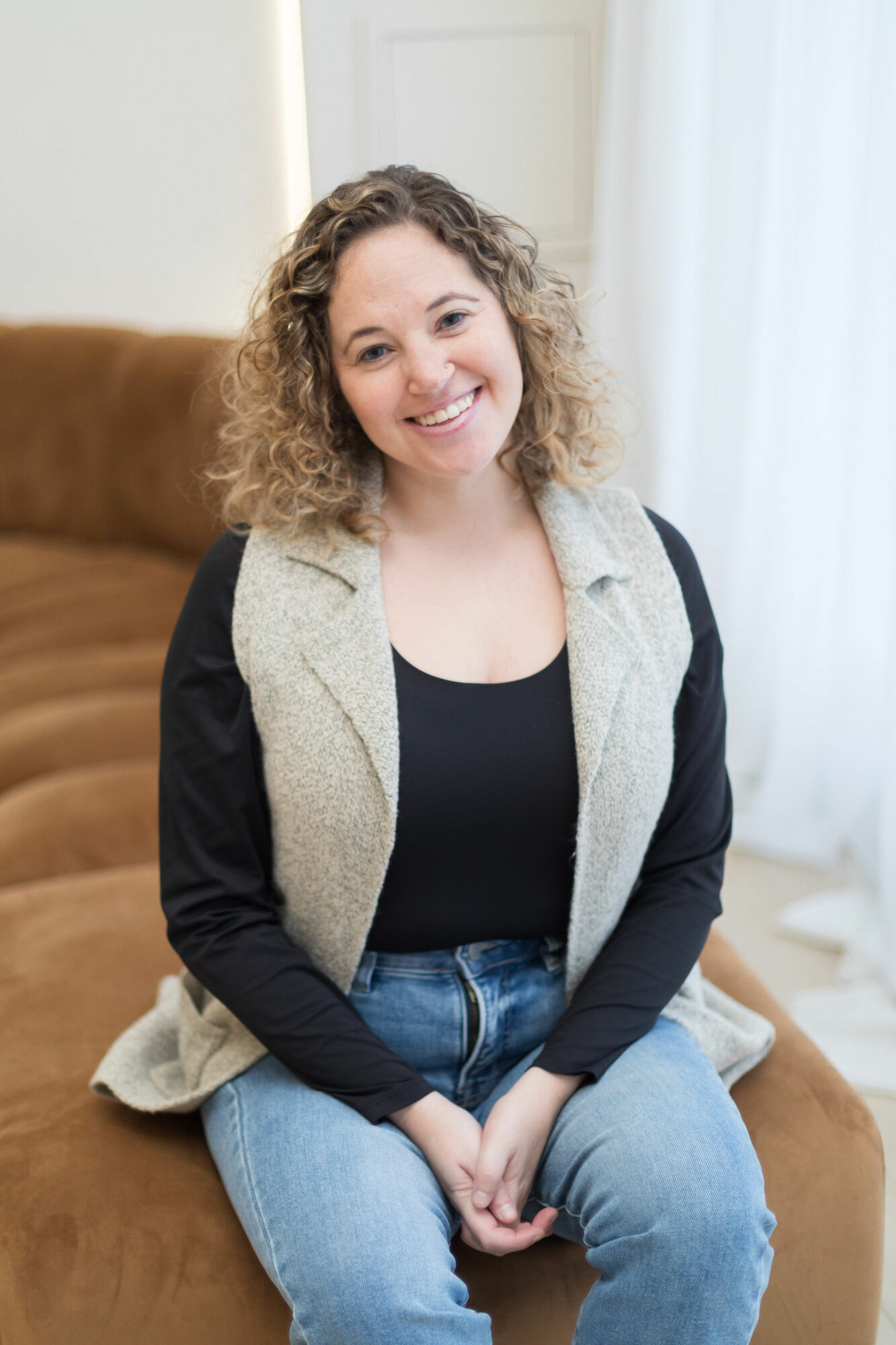 Young woman with curly hair smiling, sitting on a brown couch in a bright room with white curtains.